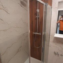 Kadek Usługi Remontowo-Wykończeniowe - Nowoczesna kabina prysznicowa z chromowaną armaturą na tle marmurowych płytek i drewnianej ściany, widoczny fragment osoby robiącej zdjęcie w lustrze.