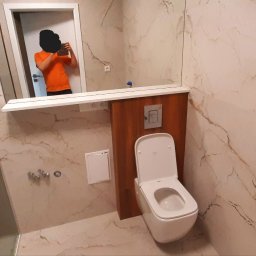 Kadek Usługi Remontowo-Wykończeniowe - Nowoczesna łazienka z marmurowymi płytkami, podwieszana toaleta z drewnianą obudową, szklana kabina prysznicowa i osoba robiąca zdjęcie w lustrze.