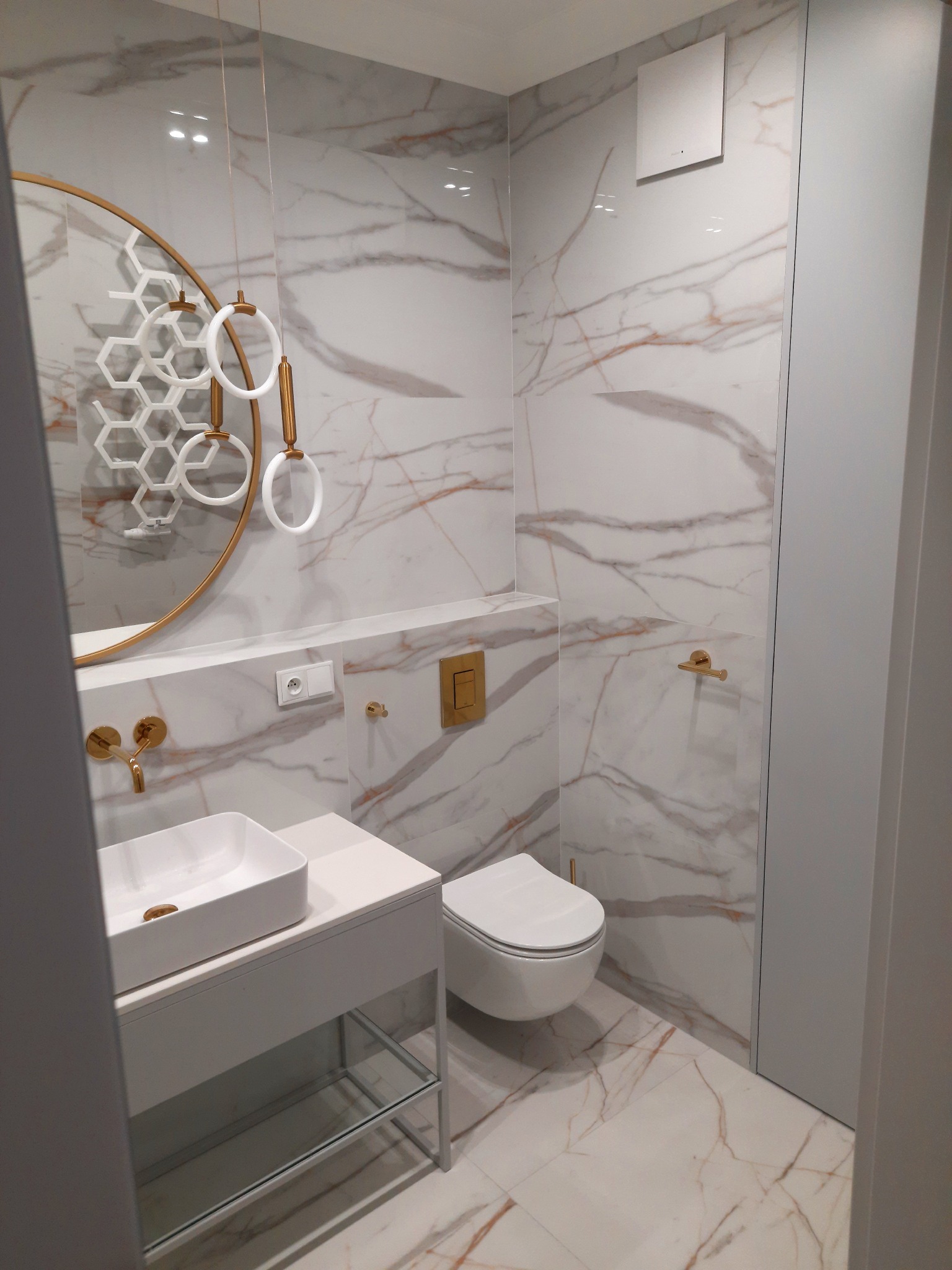 Nowoczesna łazienka w Warszawie z marmurowymi płytkami, złotymi akcentami armatury i oświetlenia, wiszącą toaletą oraz umywalką na szafce z szklaną półką.