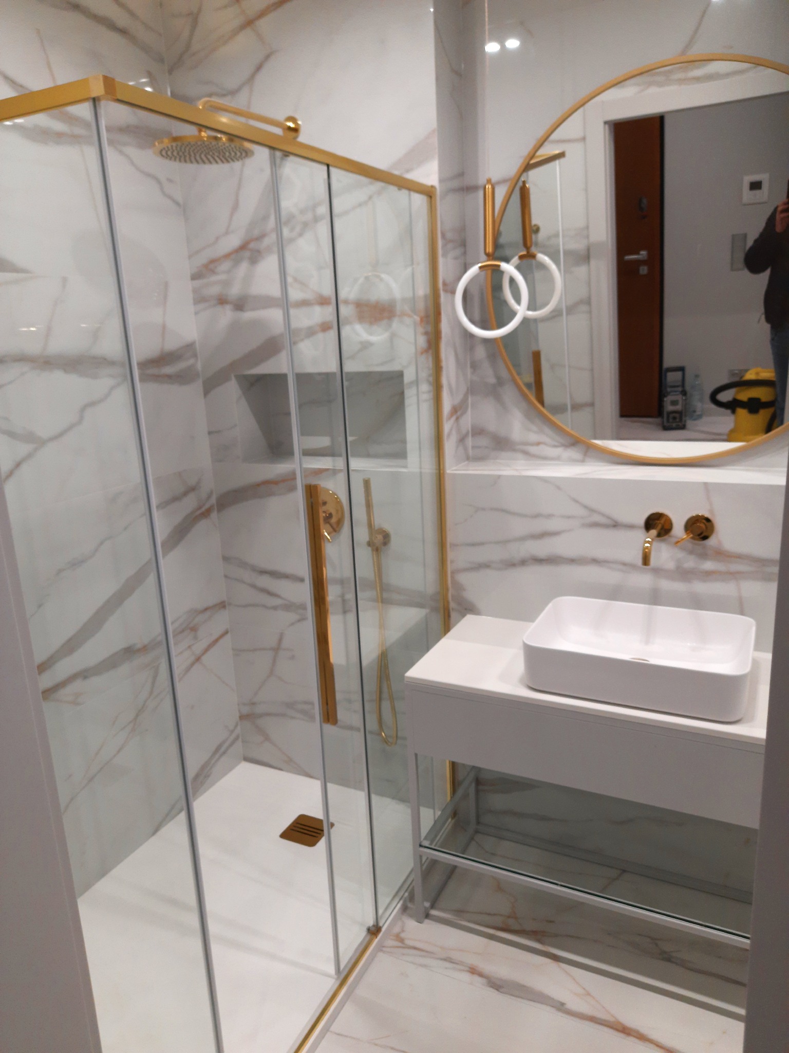 Nowoczesna łazienka z kabiną prysznicową ze złotymi ramami, białą umywalką nablatową, okrągłym lustrem i marmurowymi płytkami na ścianach i podłodze.