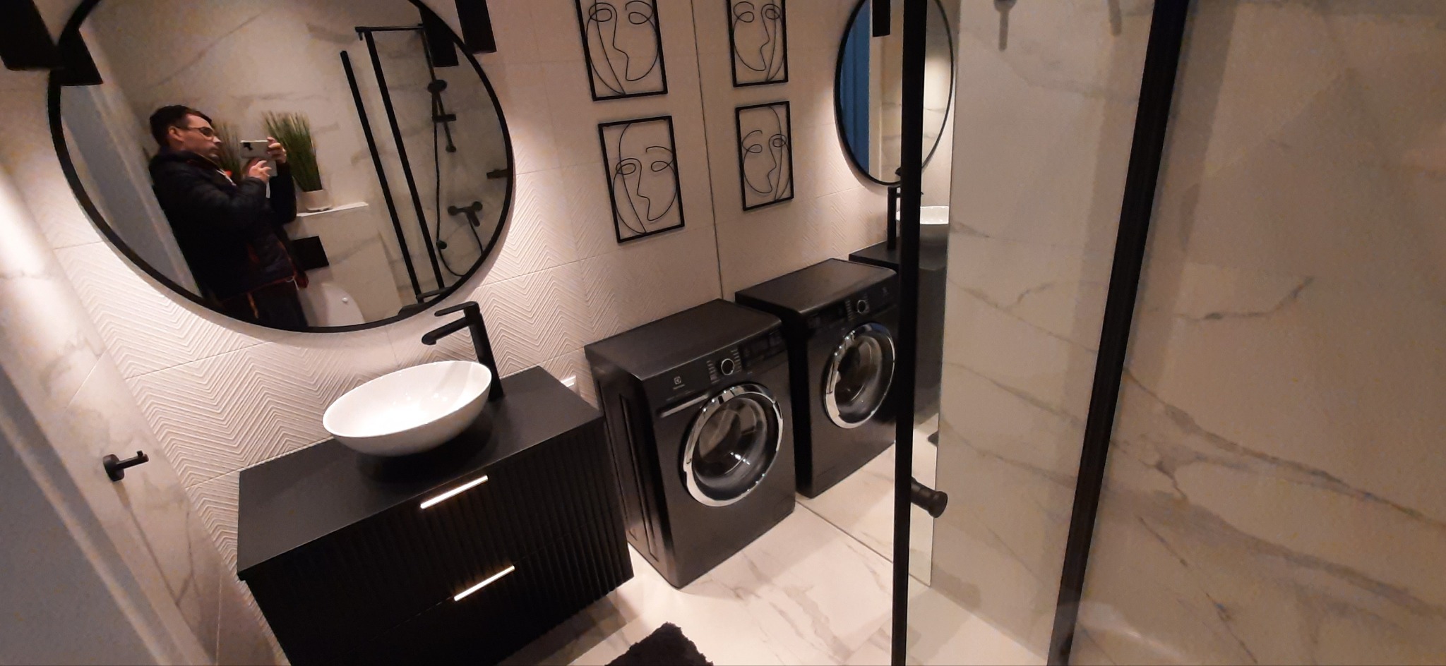 Nowoczesna łazienka z dwoma czarnymi pralkami, umywalką nablatową na czarnej szafce z frezowanymi frontami, okrągłym lustrem z odbiciem fotografa i abstrakcyjnymi obrazkami na ścianie...