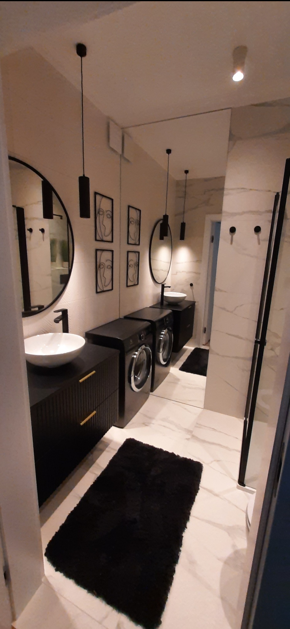 Nowoczesna łazienka z marmurową podłogą i ścianami, czarne szafki z umywalkami w kształcie misy, okrągłe lustra, czarne wiszące lampy, pralka w zabudowie meblowej oraz abstrakcyjne obrazy.