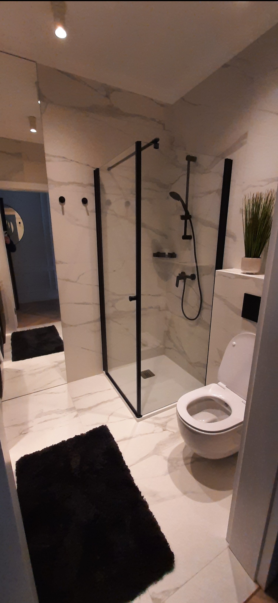 Nowoczesna łazienka z kabiną prysznicową typu walk-in z czarnymi ramami, białą toaletą, marmurowymi płytkami na ścianach i podłodze oraz czarnym dywanikiem.
