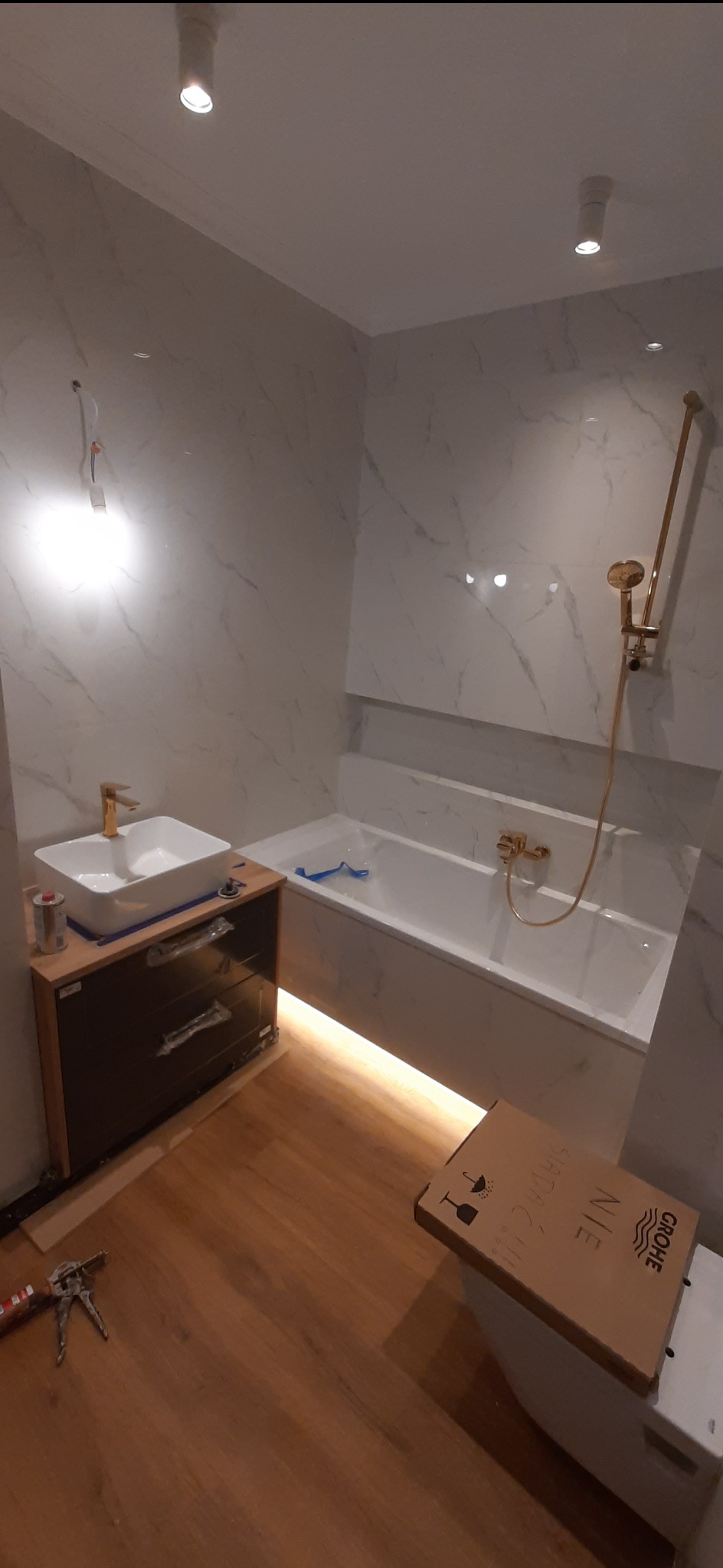 Nowoczesna łazienka w trakcie wykańczania: białe marmurowe płytki, złota armatura, podświetlana wanna, drewnopodobna podłoga, karton po armaturze na sedesie, widoczne narzędzia glazurnicze.