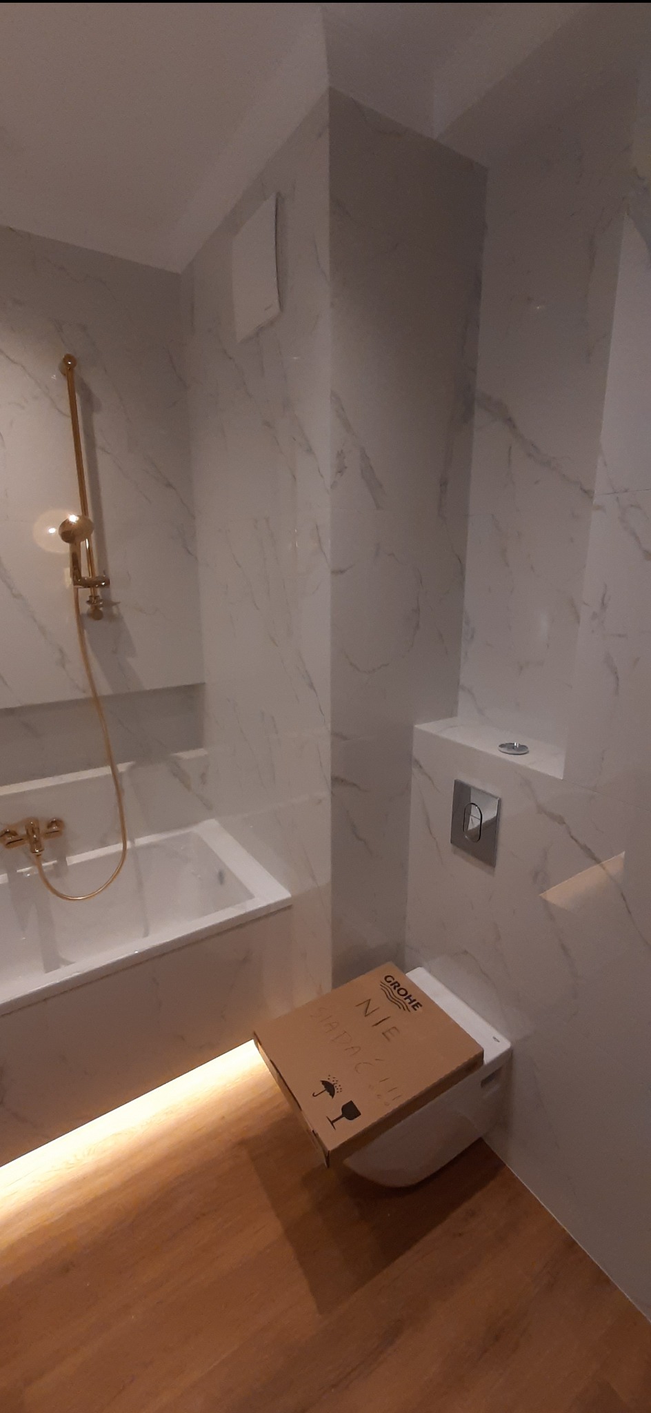 Nowoczesna łazienka z marmurowymi płytkami, złotą armaturą i podświetlaną wanną, z kartonem po nowej toalecie Grohe leżącym na podłodze.