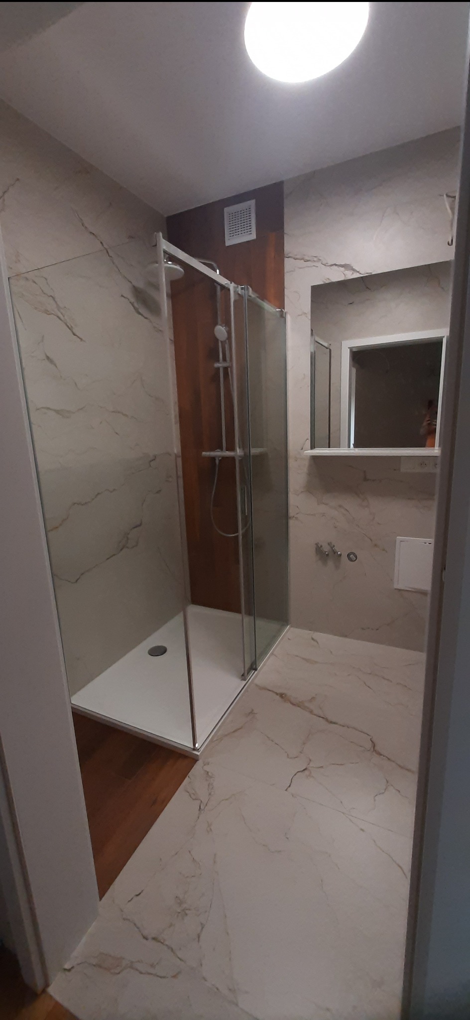 Nowoczesna łazienka z kabiną prysznicową typu walk-in, wykończona marmurowymi płytkami w odcieniach beżu i brązu, z akcentem drewnianej ściany za kabiną i drewnianą podłogą w części wejściowej.