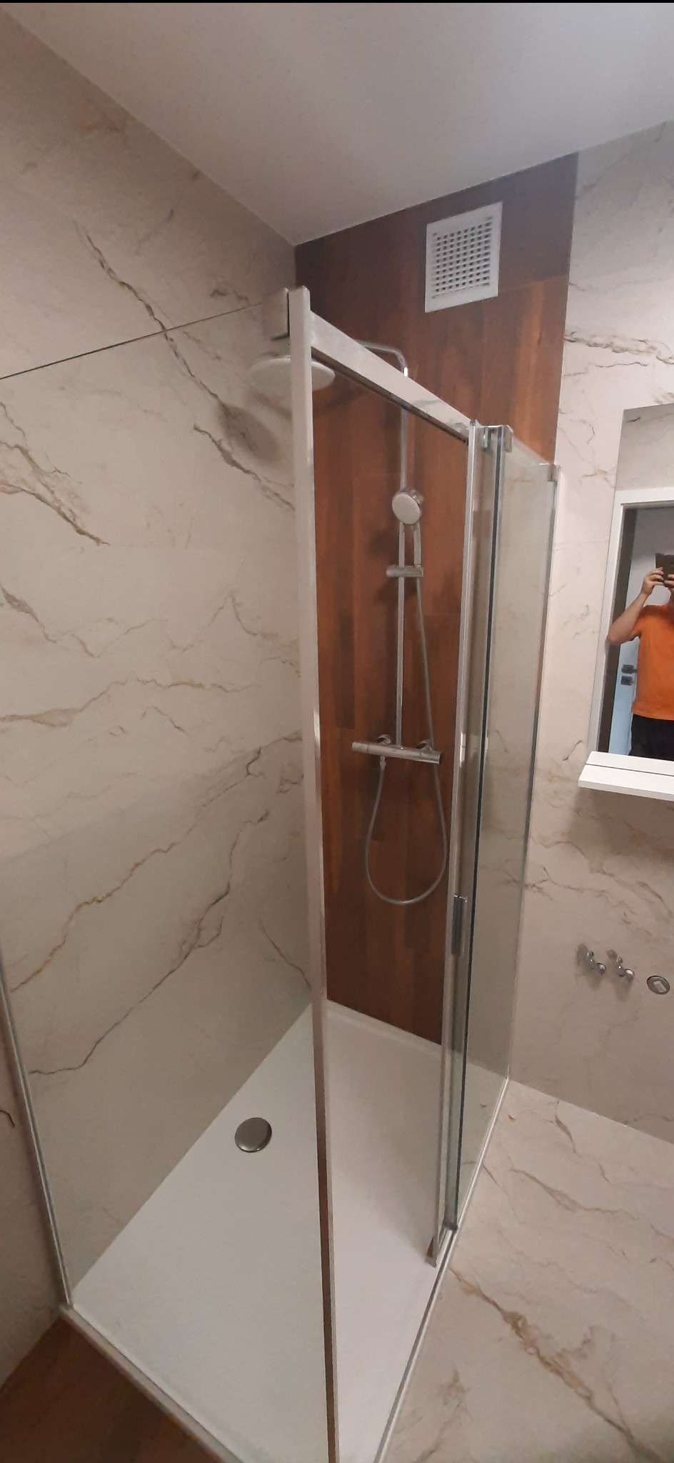 Nowoczesna kabina prysznicowa z chromowaną armaturą na tle marmurowych płytek i drewnianej ściany, widoczny fragment osoby robiącej zdjęcie w lustrze.