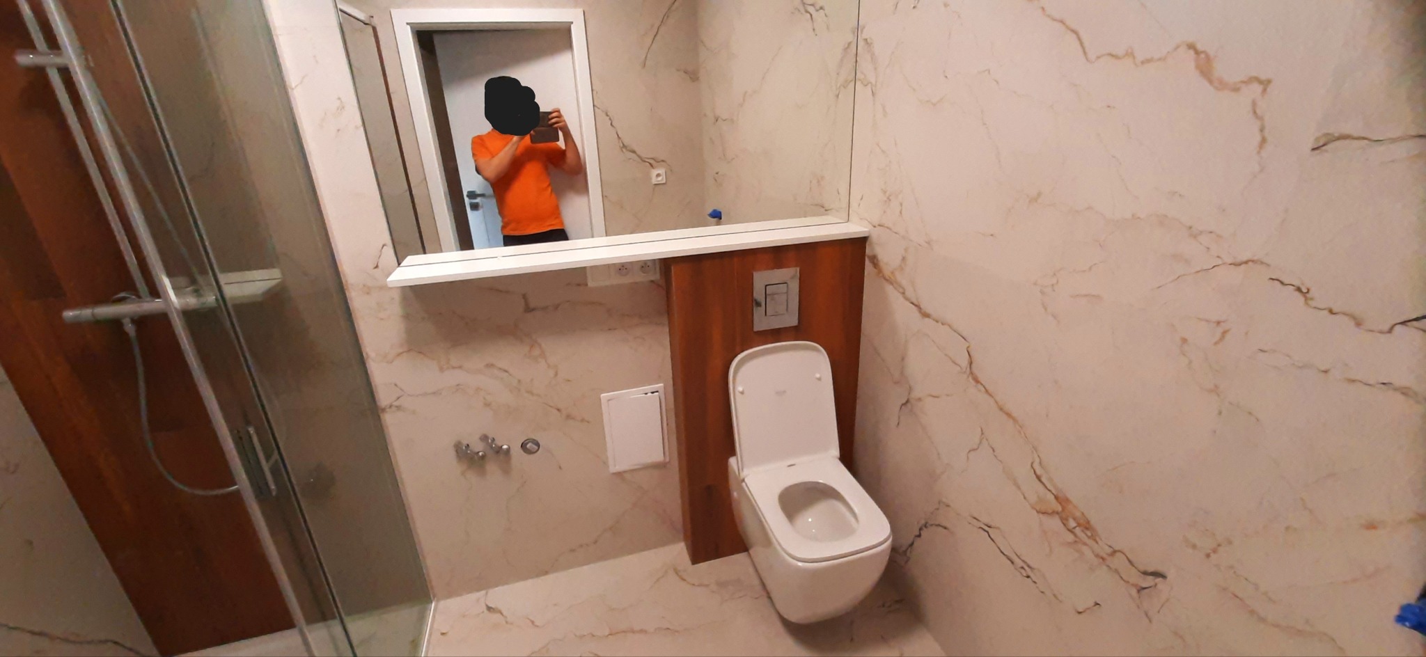 Nowoczesna łazienka z marmurowymi płytkami, podwieszana toaleta z drewnianą obudową, szklana kabina prysznicowa i osoba robiąca zdjęcie w lustrze.