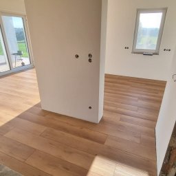 NEOWNĘTRZE SPÓŁKA Z OGRANICZONĄ ODPOWIEDZIALNOŚCIĄ - Jasne, drewnopodobne płytki podłogowe ułożone w nowym, pustym pomieszczeniu z dużym oknem balkonowym i widokiem na zewnątrz. Ściany białe, widać niezabezpieczone otwory na gniazdka elektryczne.