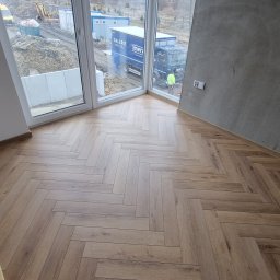 NEOWNĘTRZE SPÓŁKA Z OGRANICZONĄ ODPOWIEDZIALNOŚCIĄ - Ułożona podłoga z paneli w jodełkę, widok na okno z placem budowy i zaparkowany samochód dostawczy.