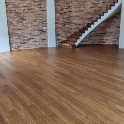 NEOWNĘTRZE SPÓŁKA Z OGRANICZONĄ ODPOWIEDZIALNOŚCIĄ - Ułożona, brązowa podłoga drewniana w przestronnym wnętrzu z ceglaną ścianą i drewnianymi schodami, widoczne pionowe rury instalacyjne.