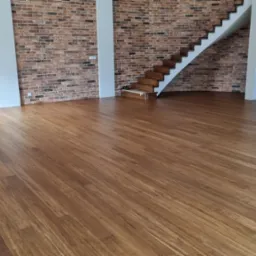 Ułożona, brązowa podłoga drewniana w przestronnym wnętrzu z ceglaną ścianą i drewnianymi schodami, widoczne pionowe rury instalacyjne.