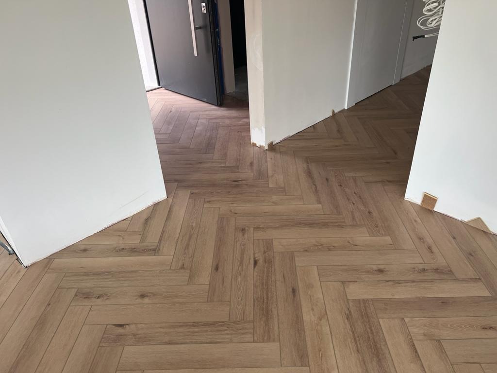 Ułożona podłoga z paneli w jodełkę w jasnym odcieniu drewna, widoczne niedokończone wykończenia przy ścianach i otworach drzwiowych.