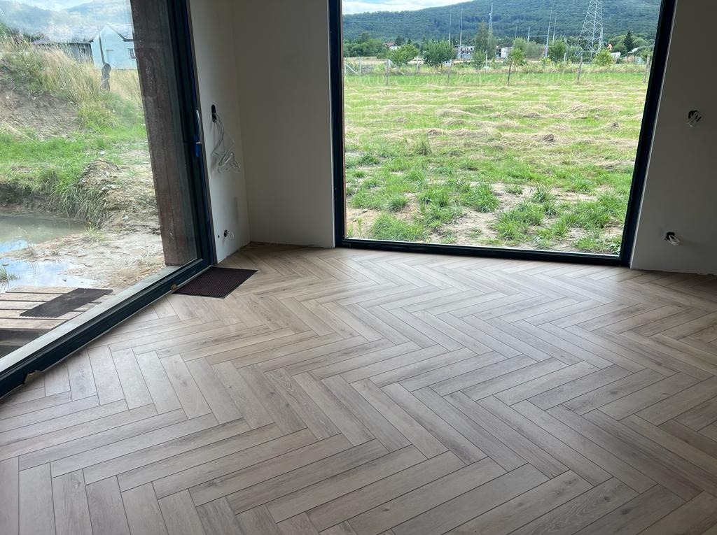 Jasne panele ułożone we wzór jodełki francuskiej w nowym pomieszczeniu z widokiem na zewnątrz przez duże okna.