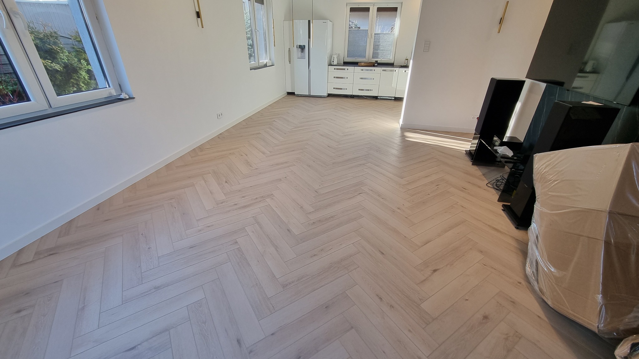 Jasne panele ułożone w jodełkę francuską w przestronnym salonie z oknami i białymi meblami kuchennymi w tle, obok zapakowanych głośników.