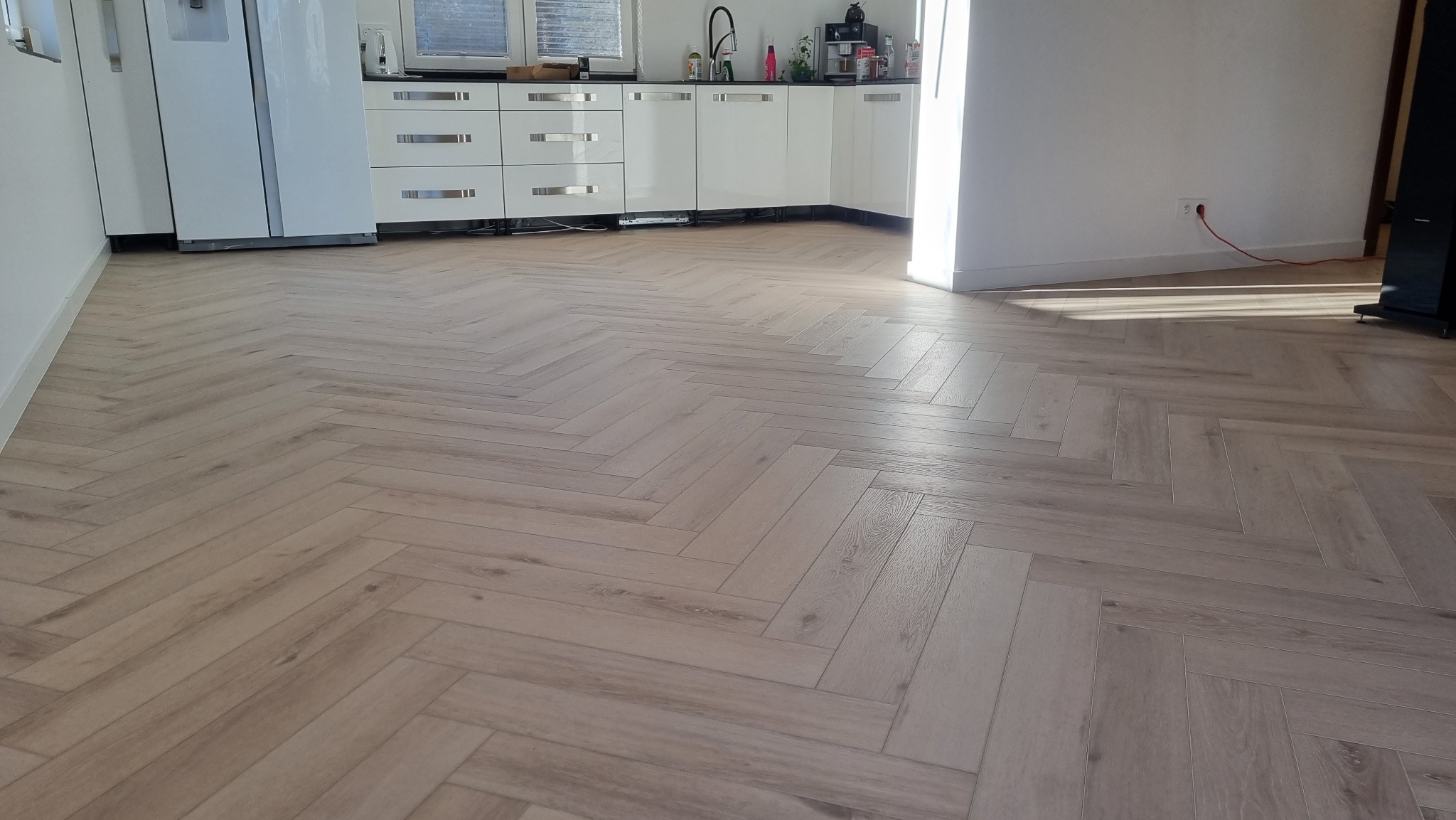 Jasna, drewnopodobna podłoga ułożona we wzór jodełki francuskiej w nowoczesnej kuchni z białymi meblami.