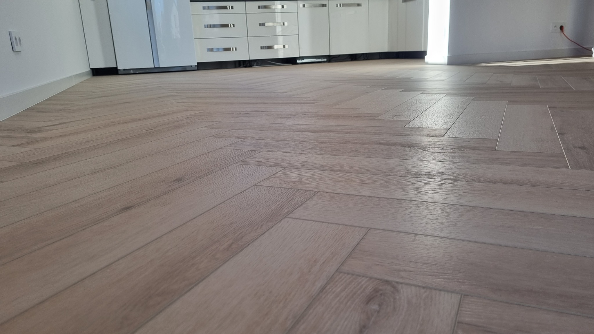 Ułożona w jodełkę jasna podłoga z paneli w nowym mieszkaniu, widok z niskiej perspektywy na tle białych mebli kuchennych.