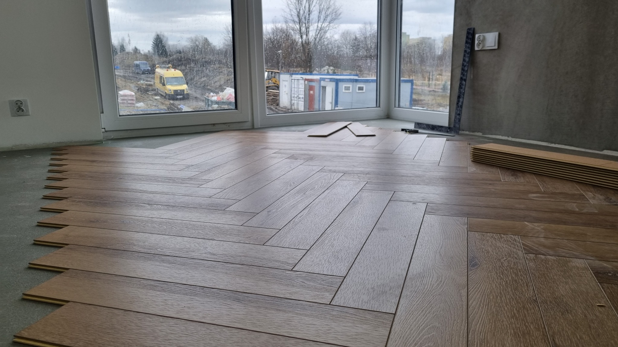 Układanie paneli w jodełkę w nowym budownictwie, widoczne narzędzia pomiarowe przy ścianie, przez okno widok na plac budowy i zaparkowany żółty bus.