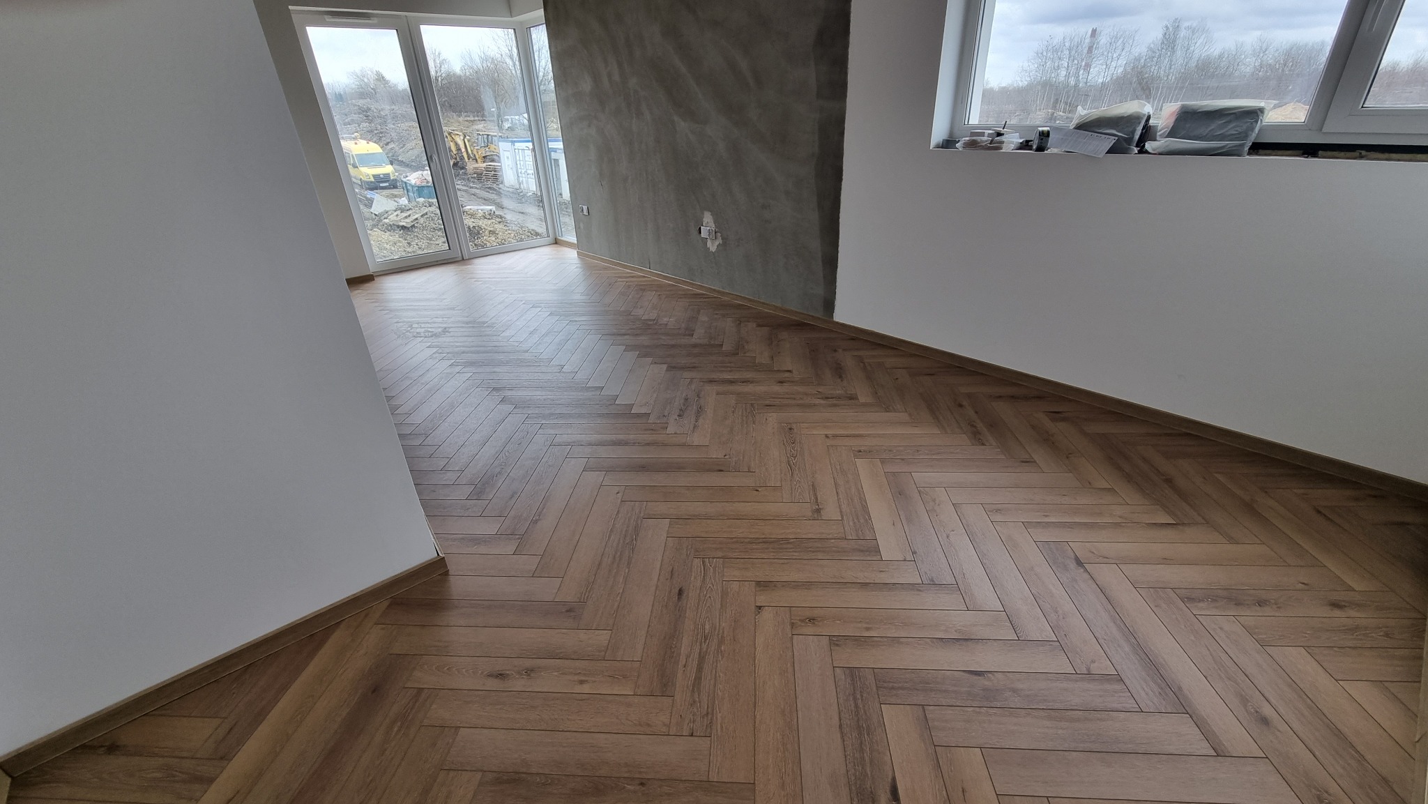 Ułożona podłoga z paneli w jodełkę, widok na budowę przez okno, surowy beton na ścianie.