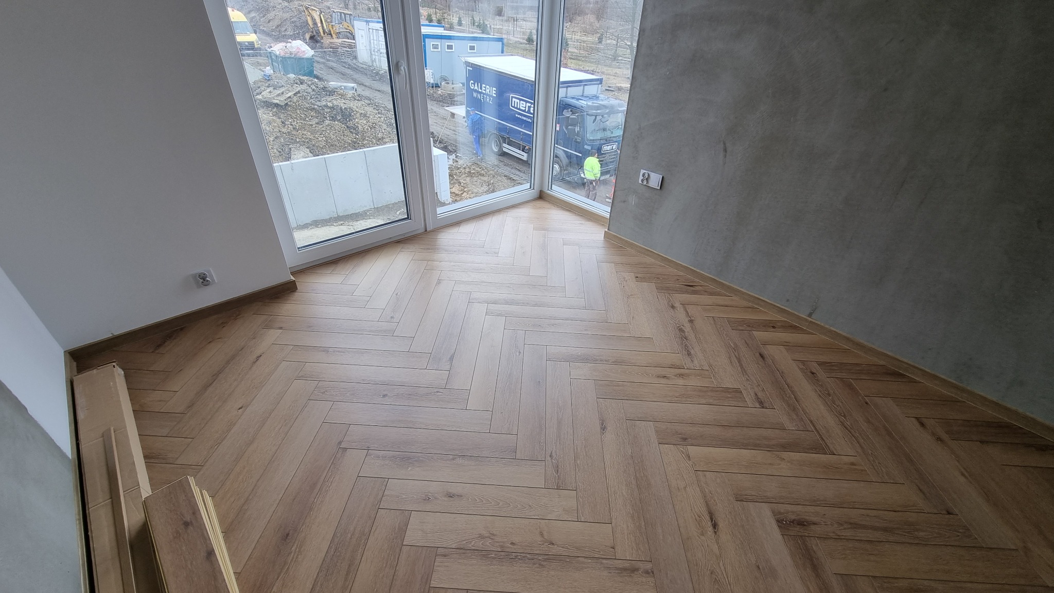 Ułożona podłoga z paneli w jodełkę, widok na okno z placem budowy i zaparkowany samochód dostawczy.