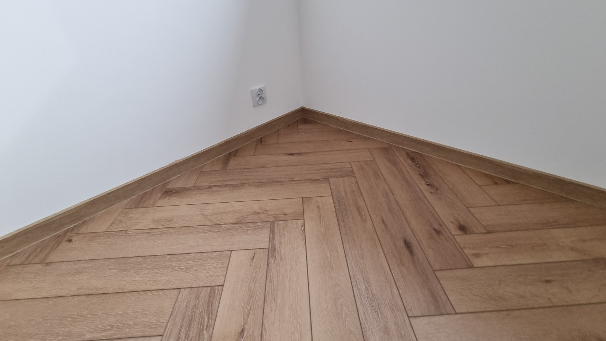 Ukończona podłoga z paneli ułożonych we wzór jodełki, widok z narożnika pomieszczenia, listwa przypodłogowa w kolorze paneli.