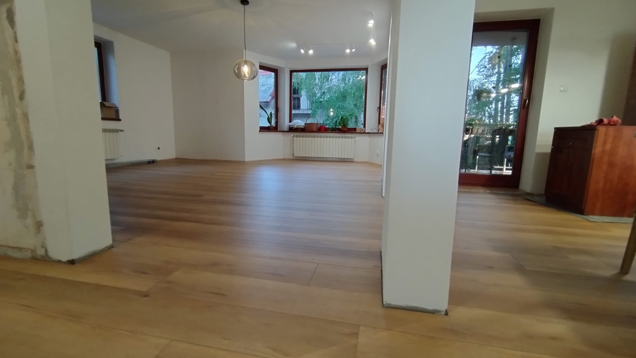 Jasna, drewniana podłoga w przestronnym salonie z dużym oknem wykuszowym i widokiem na zieleń, z kolumną w centrum kadru i wiszącą lampą.