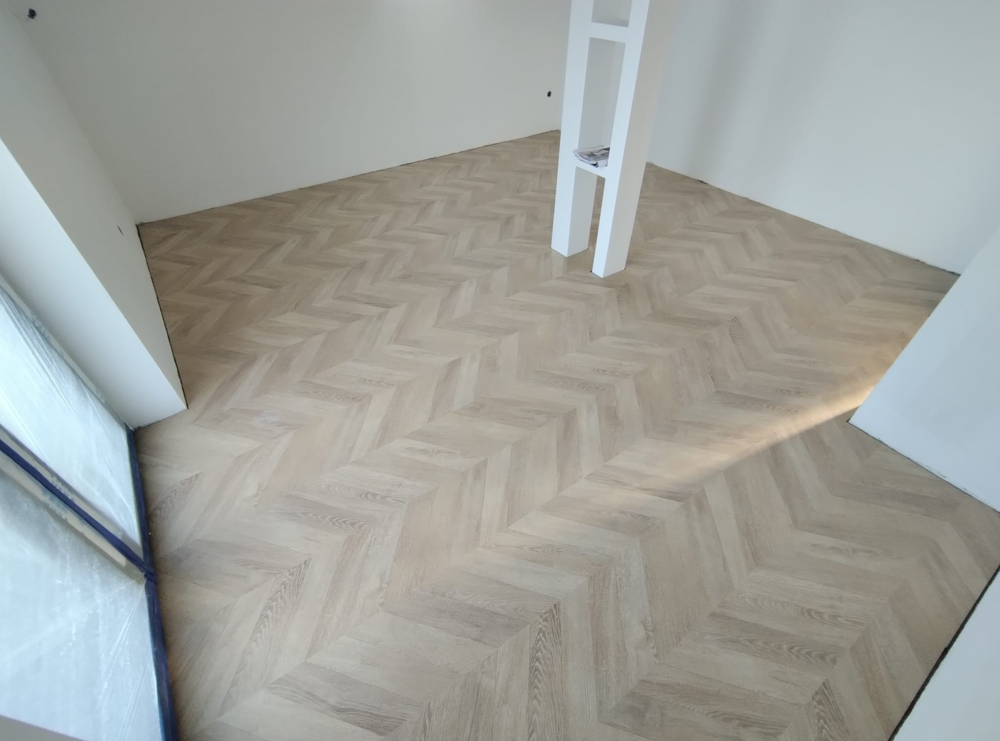 Jasne panele ułożone w jodełkę francuską w nowym, jeszcze pustym pomieszczeniu z białą ścianą i wbudowaną półką.