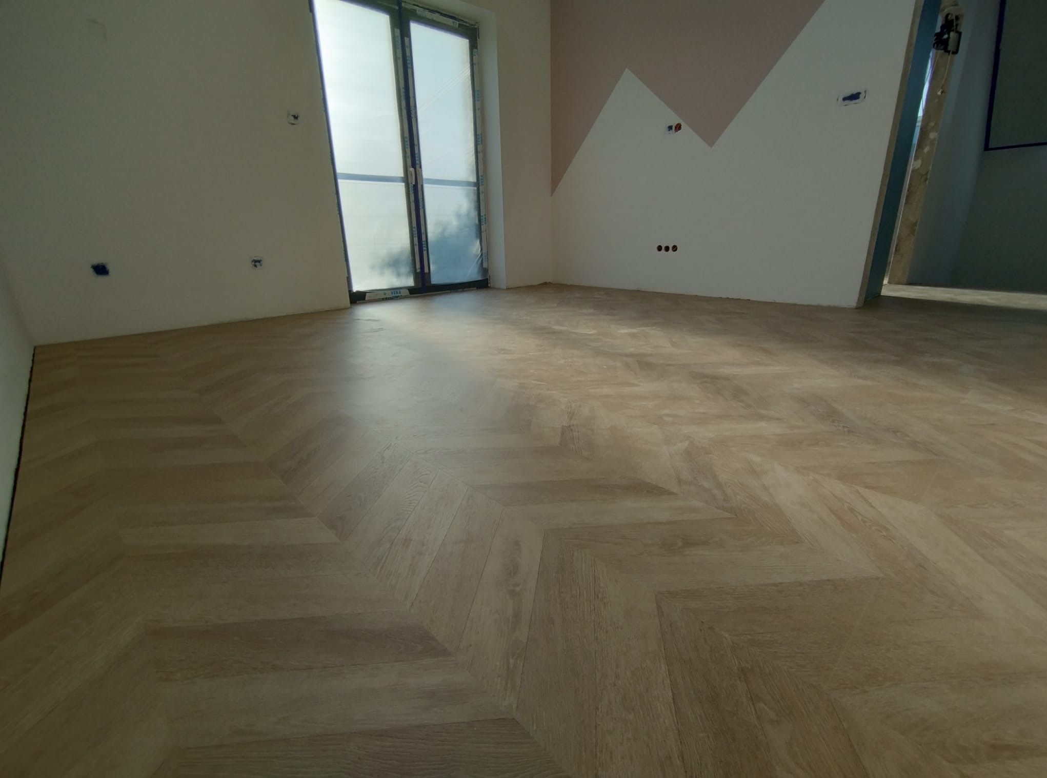 Ułożona podłoga z paneli w jodełkę w jasnym odcieniu drewna, widok na okno zabezpieczone folią i fragment ściany z geometrycznym malowaniem w kolorze różowym.