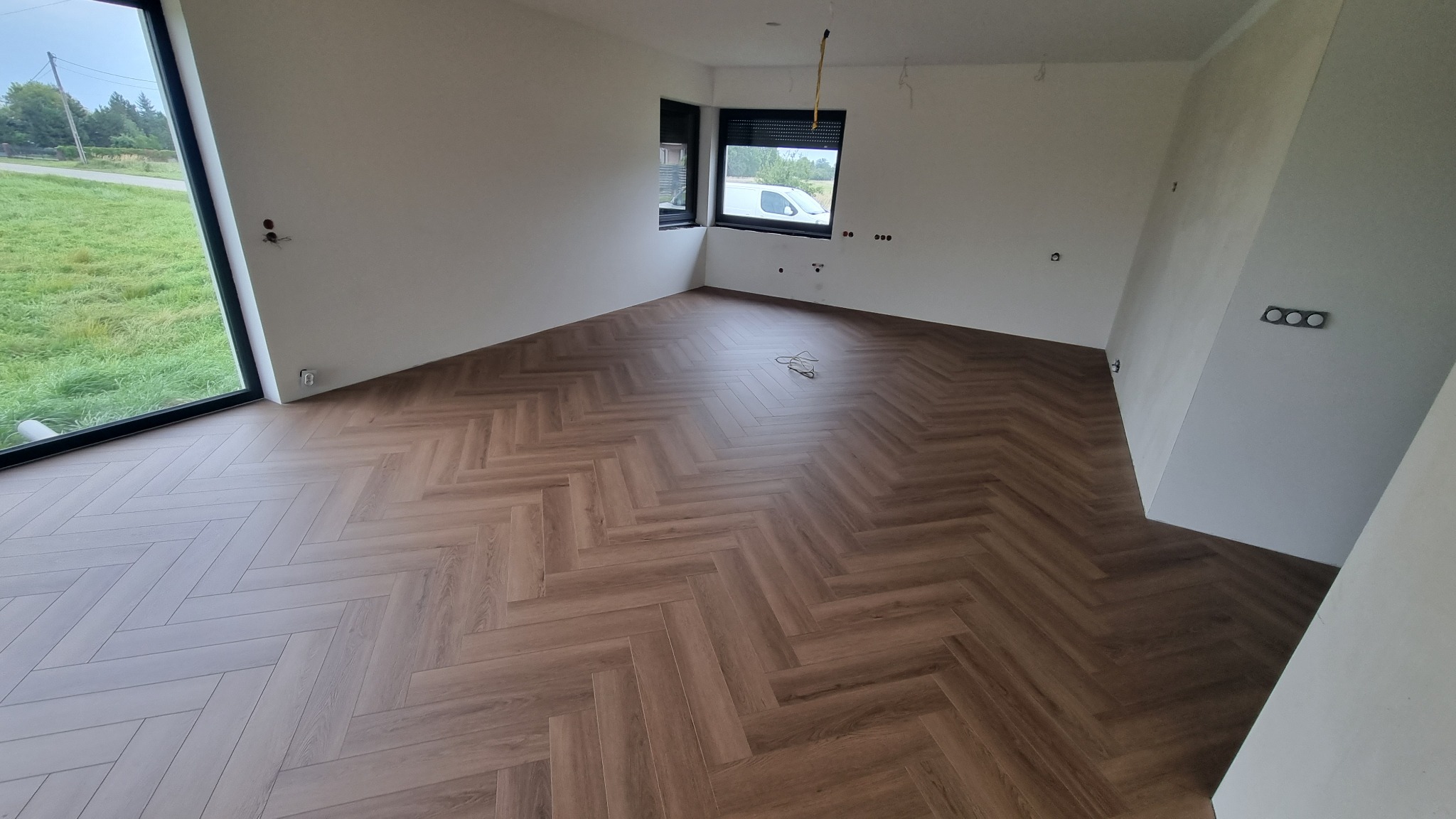 Nowo ułożona podłoga z paneli w jodełkę w jasnym, nowoczesnym wnętrzu. Widoczne gniazdka elektryczne na ścianach i przewody na podłodze, sugerujące prace wykończeniowe.