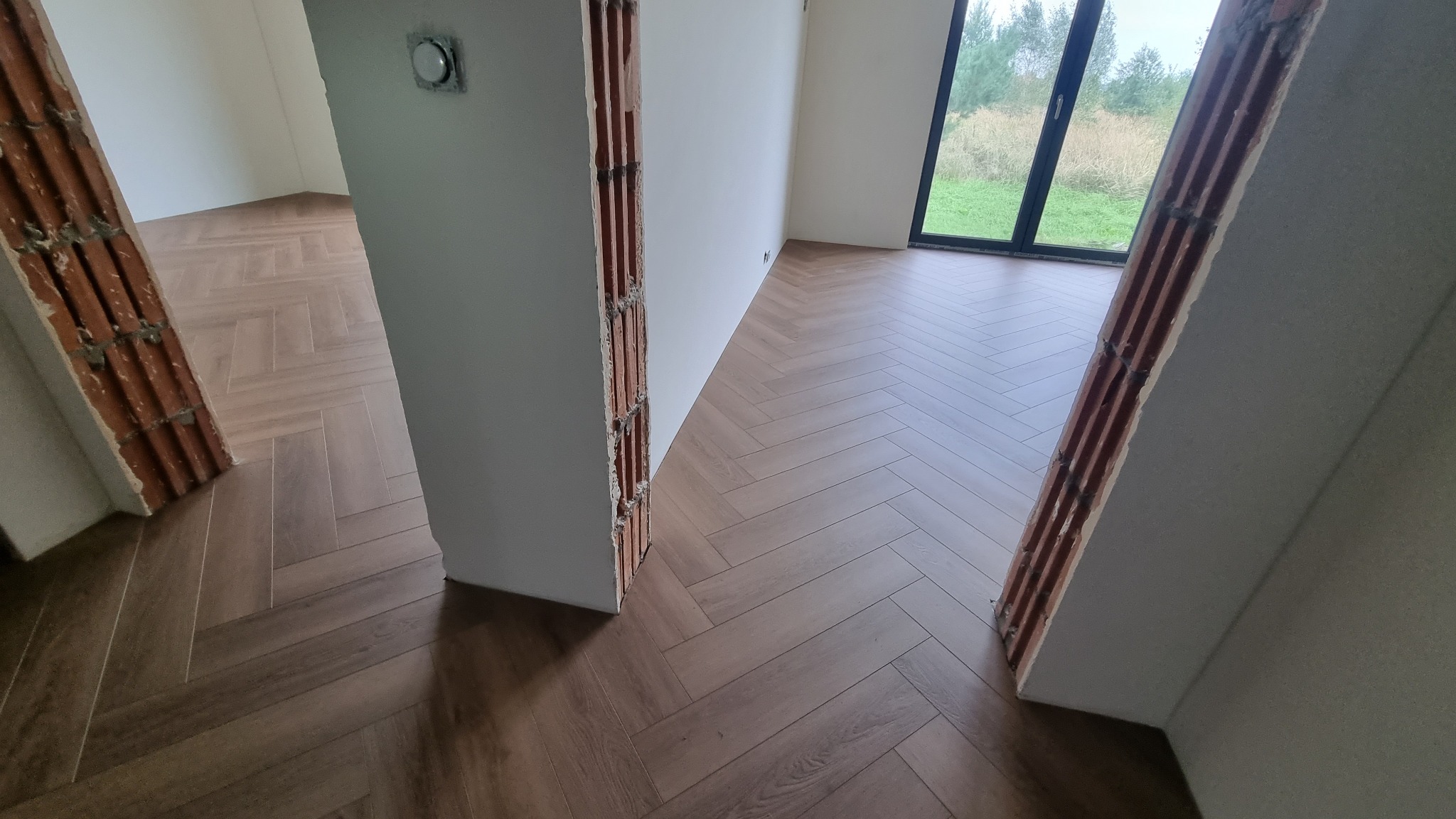 Ułożona podłoga z paneli w jodełkę, widoczne surowe wykończenia ścian i framug, okno z widokiem na zewnątrz.