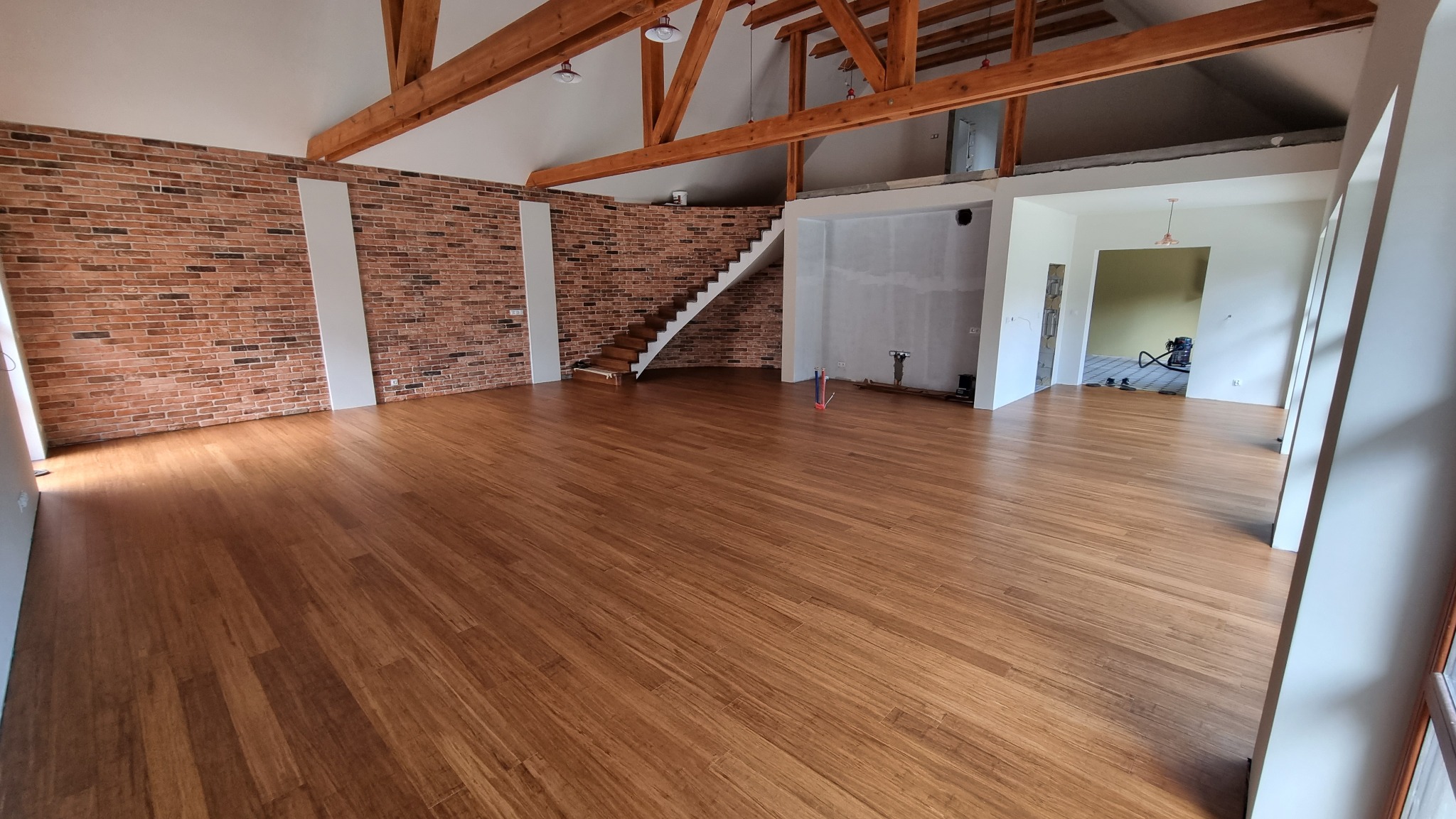 Jasny, przestronny pokój z nową, brązową podłogą z paneli, ceglana ściana z białymi wstawkami, drewniane schody i odsłonięte belki stropowe.