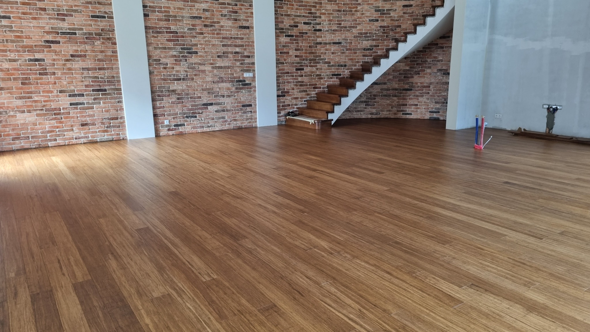 Ułożona, brązowa podłoga drewniana w przestronnym wnętrzu z ceglaną ścianą i drewnianymi schodami, widoczne pionowe rury instalacyjne.