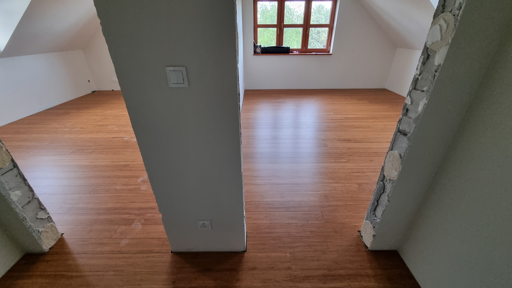 Poddasze z nową, brązową podłogą z paneli, widoczne niedokończone obramowania filarów i okno z widokiem na zieleń.