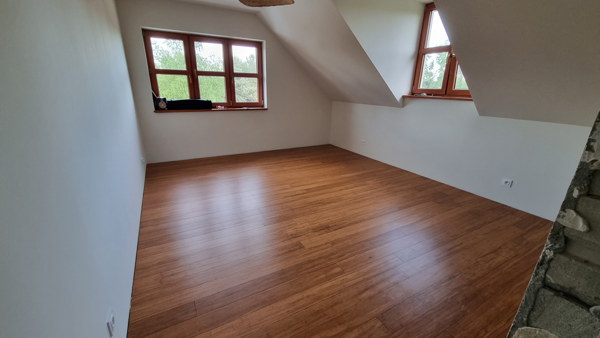 Pokój na poddaszu z oknami i świeżo ułożoną, błyszczącą podłogą z paneli w odcieniach brązu. Fragment ściany z surowej cegły w prawym dolnym rogu.