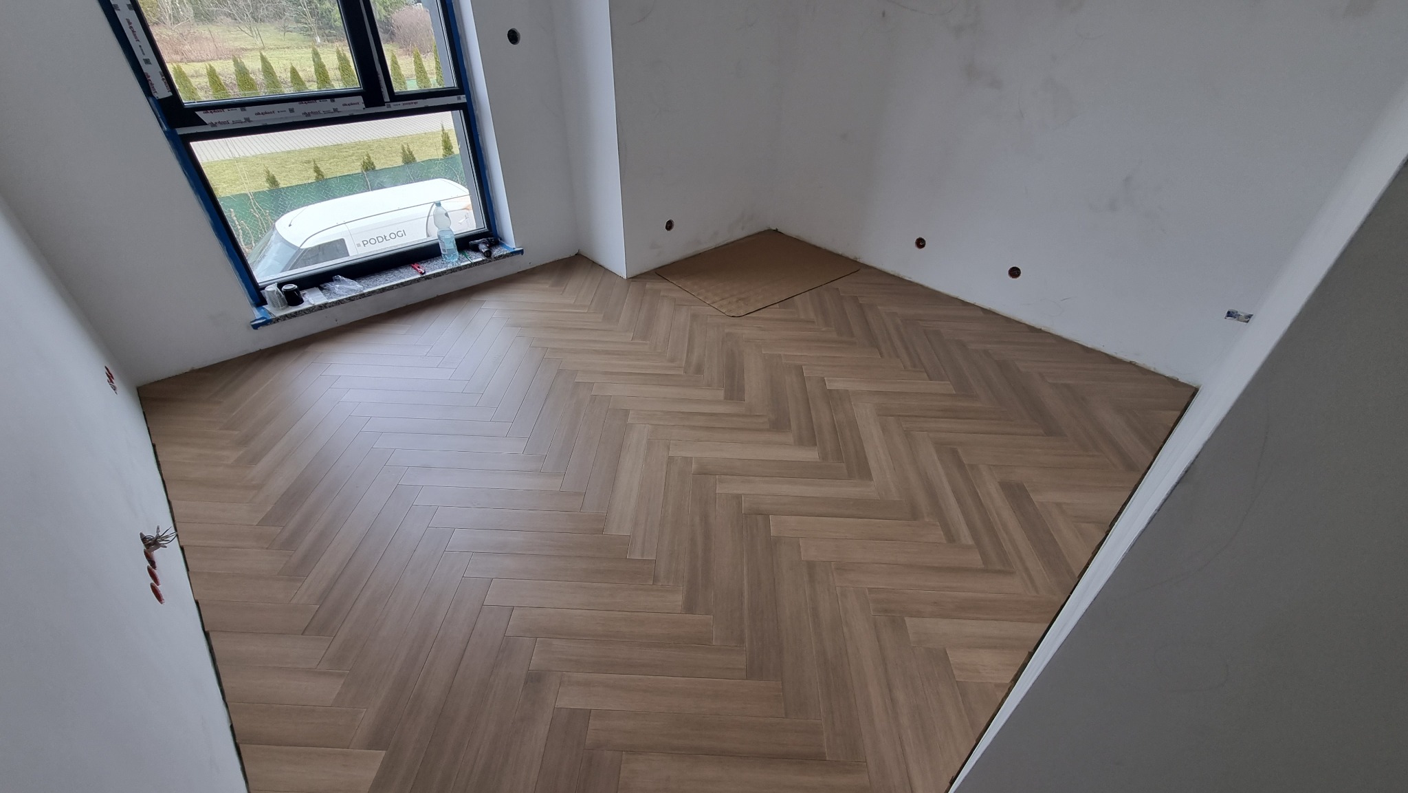 Układanie parkietu w jodełkę w jasnym pomieszczeniu z oknem, widoczne niedokończone ściany i zakryty otwór w podłodze.