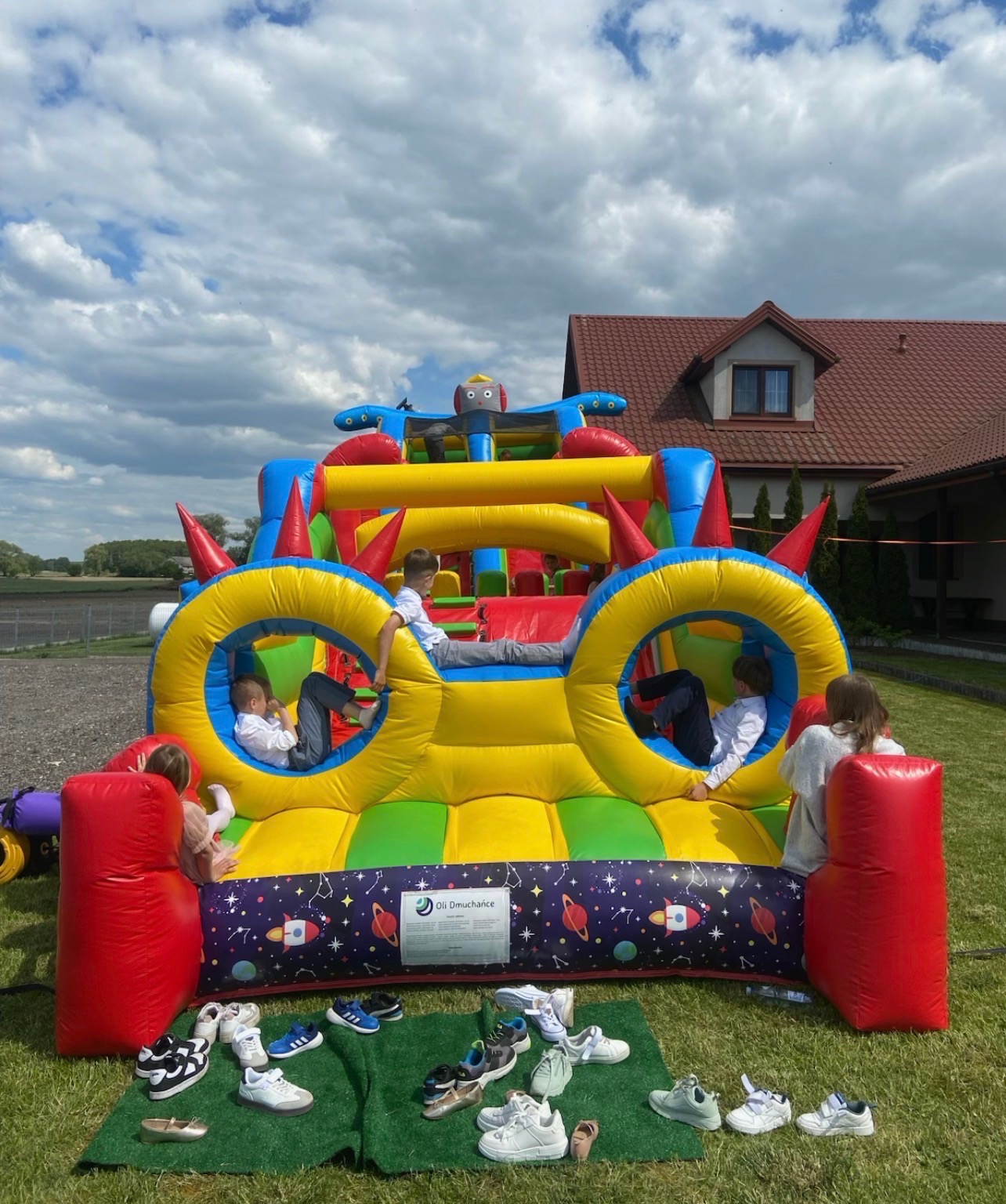 Kolorowy dmuchany zamek w kształcie pojazdu kosmicznego, dzieci bawią się w środku, buty zdjęte przed wejściem, trawnik, dom w tle, słoneczny dzień.