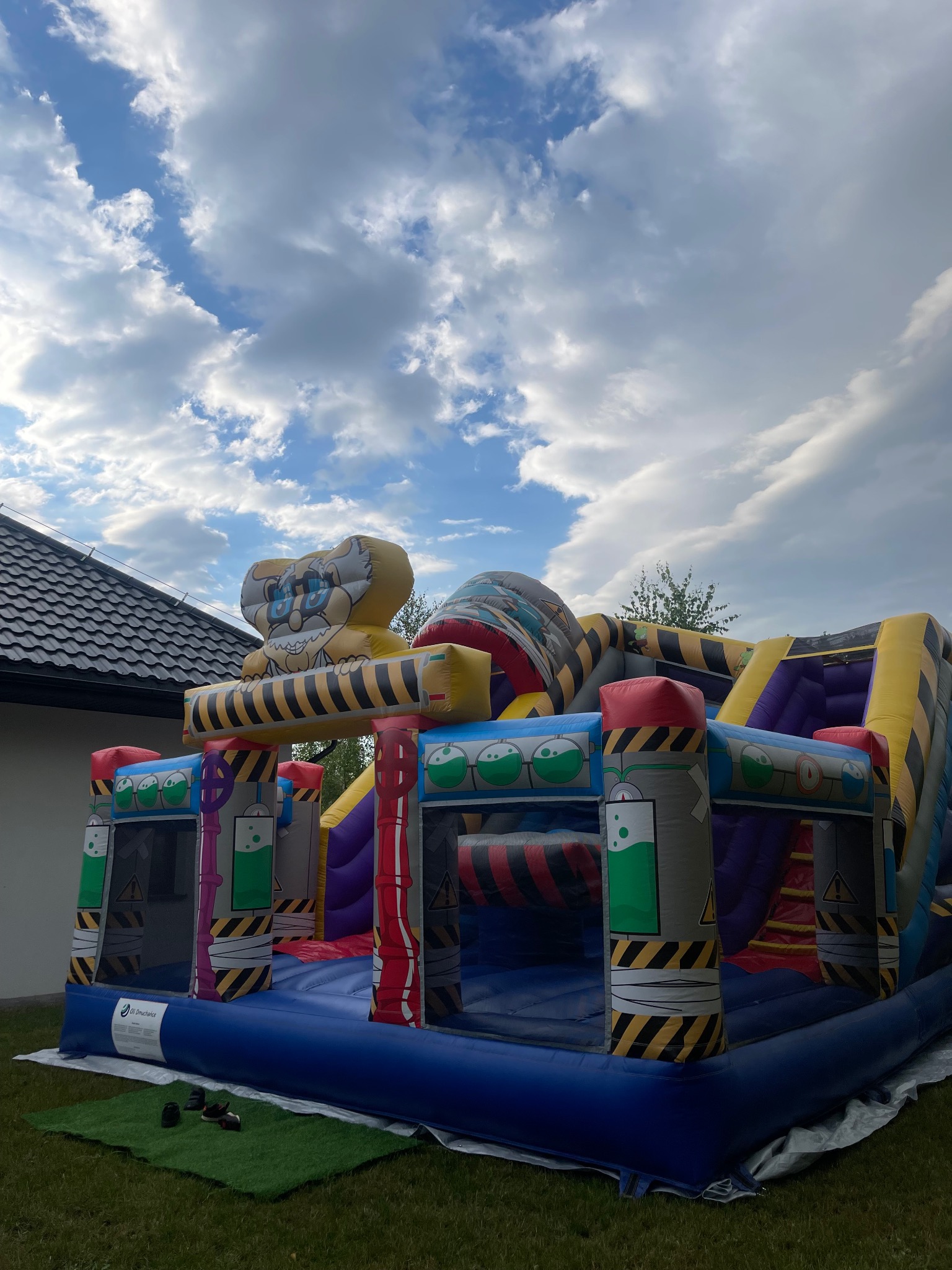 Kolorowy, dmuchany zamek z motywem laboratorium i postacią kreskówkową na szczycie, ustawiony na trawie przed domem, pod błękitnym niebem z chmurami.