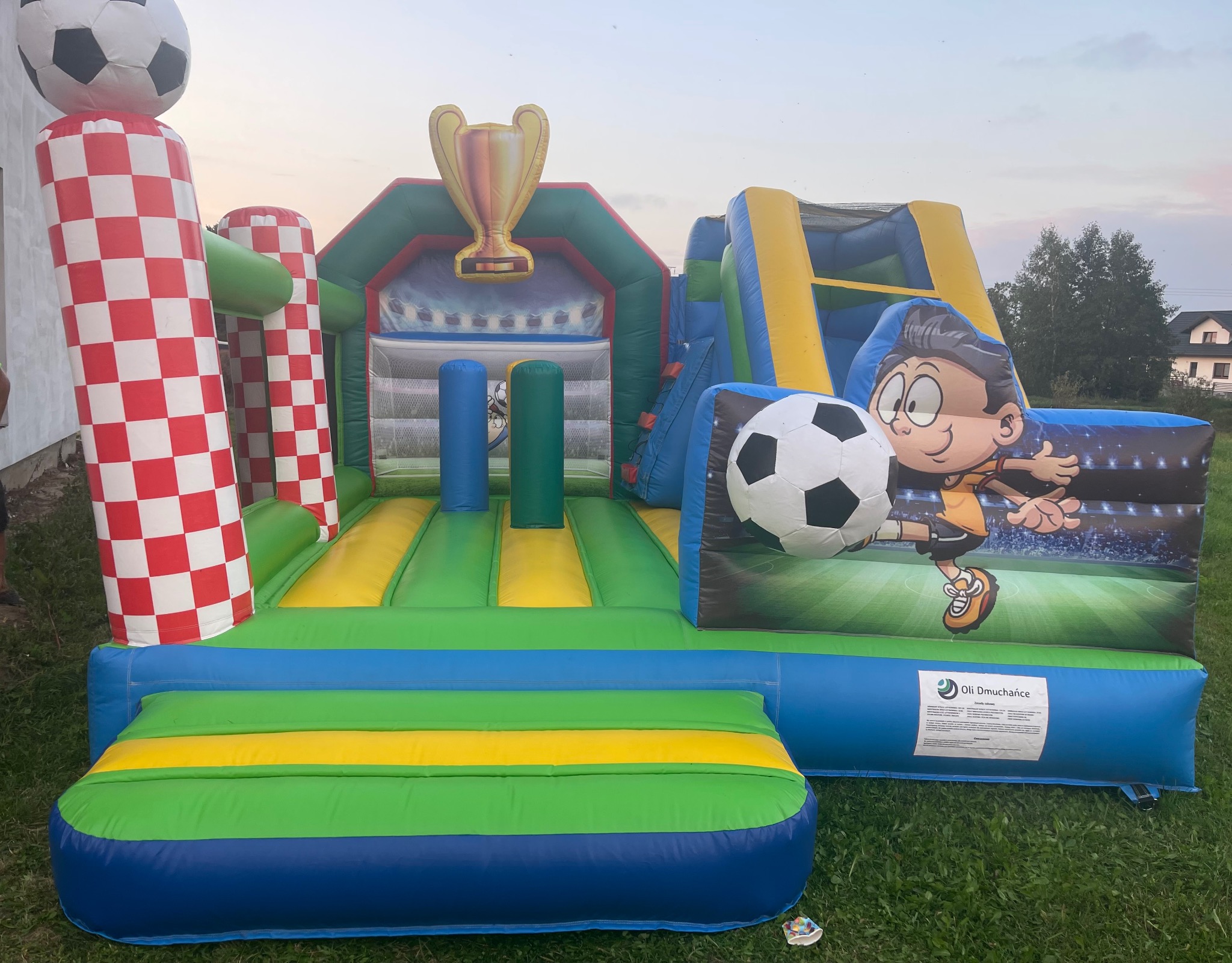 Kolorowy dmuchany zamek w tematyce piłkarskiej z grafiką chłopca z piłką i trofeum na szczycie, ustawiony na trawie. Widoczne elementy w barwach zieleni, żółci i niebieskiego.