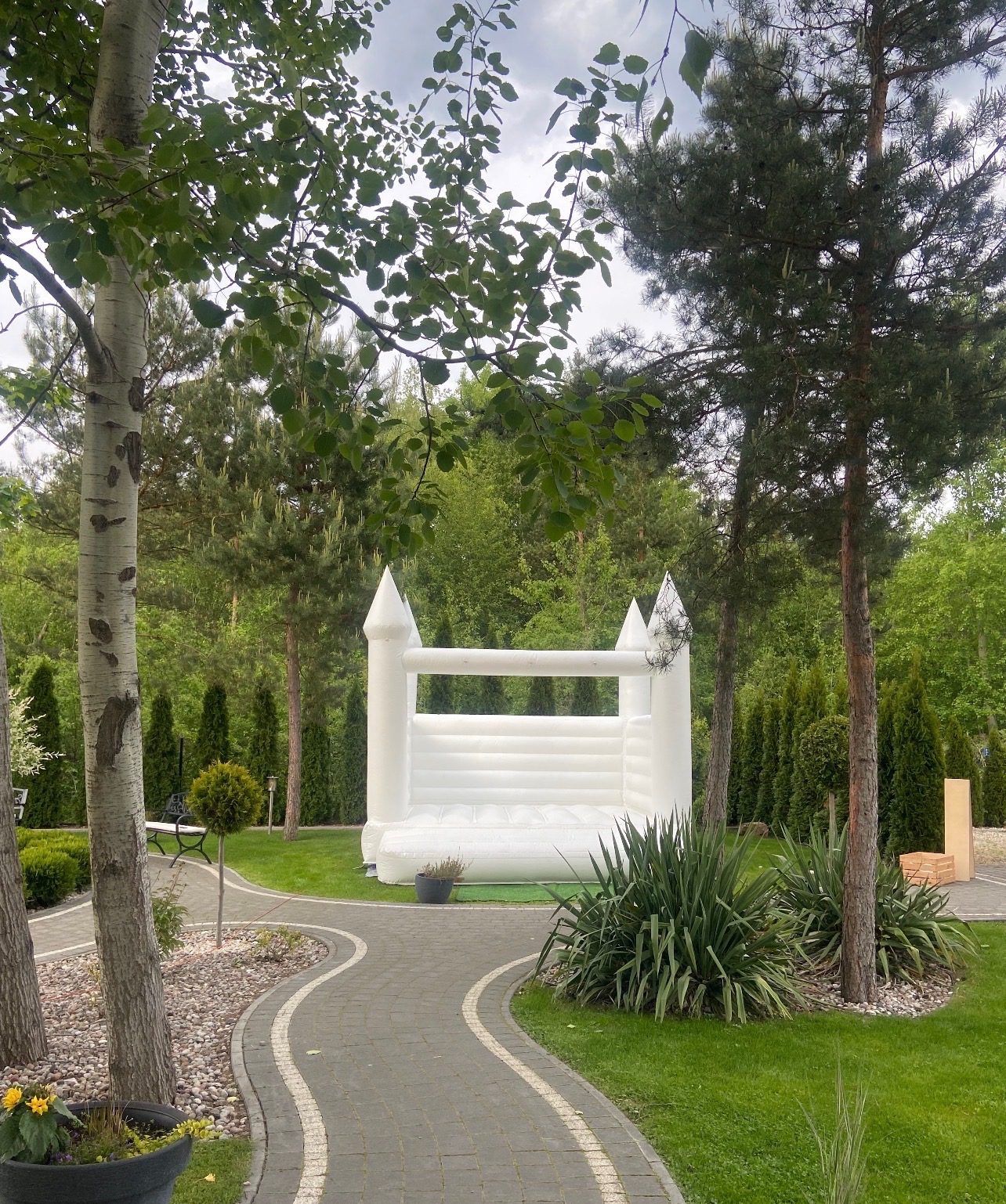 Biały dmuchany zamek na trawie, otoczony drzewami i krzewami. Kręta ścieżka prowadzi do zamku. Ujęcie z perspektywy ogrodu, w tle zieleń.