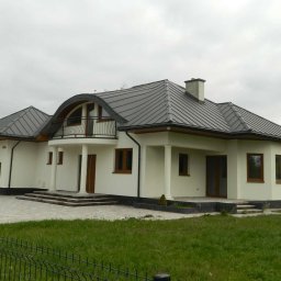 Mirosław JOŃCA MJ PROJECT - Nowoczesny, parterowy dom z grafitowym dachem blaszanym, białymi ścianami i kolumnami podtrzymującymi balkon, widoczny zza czarnego, metalowego ogrodzenia na tle pochmurnego nieba.