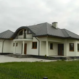 Nowoczesny, parterowy dom z grafitowym dachem blaszanym, białymi ścianami i kolumnami podtrzymującymi balkon, widoczny zza czarnego, metalowego ogrodzenia na tle pochmurnego nieba.