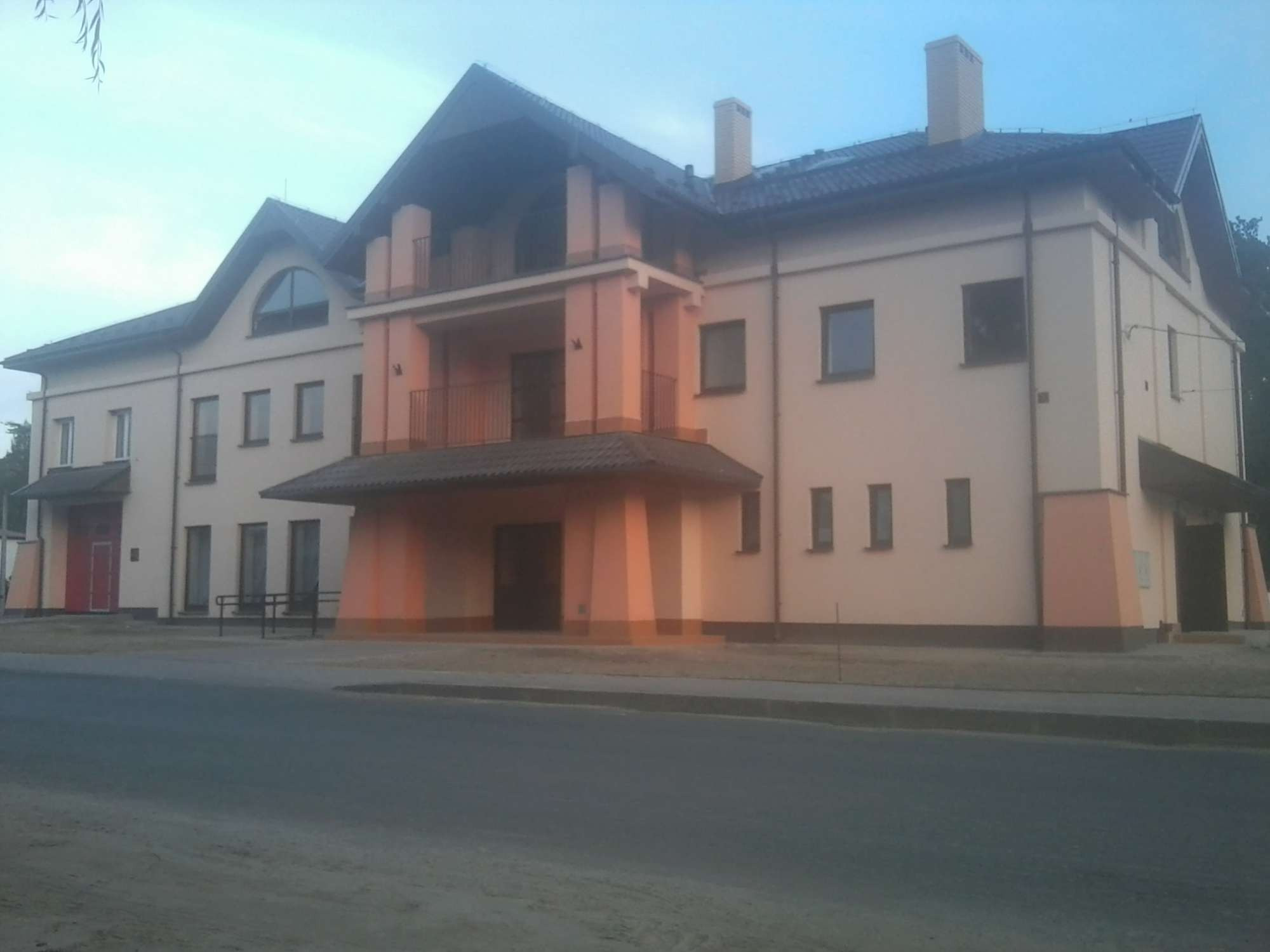 Dwupiętrowy budynek mieszkalny z balkonem i dwoma kominami, widziany z ulicy w pochmurny dzień.