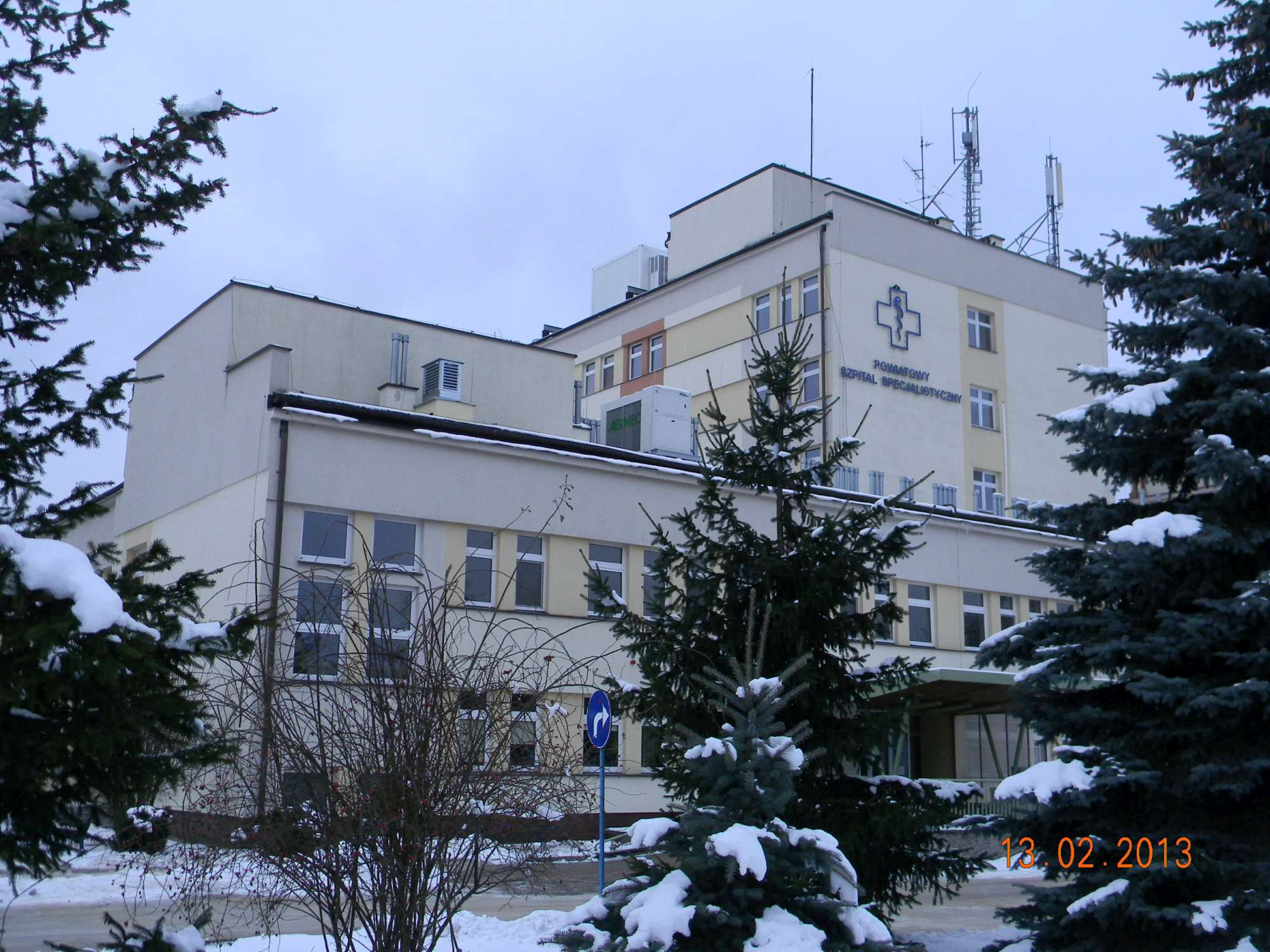 Budynek szpitala specjalistycznego w zimowej scenerii, z widocznymi antenami na dachu i drzewami iglastymi pokrytymi śniegiem na pierwszym planie.
