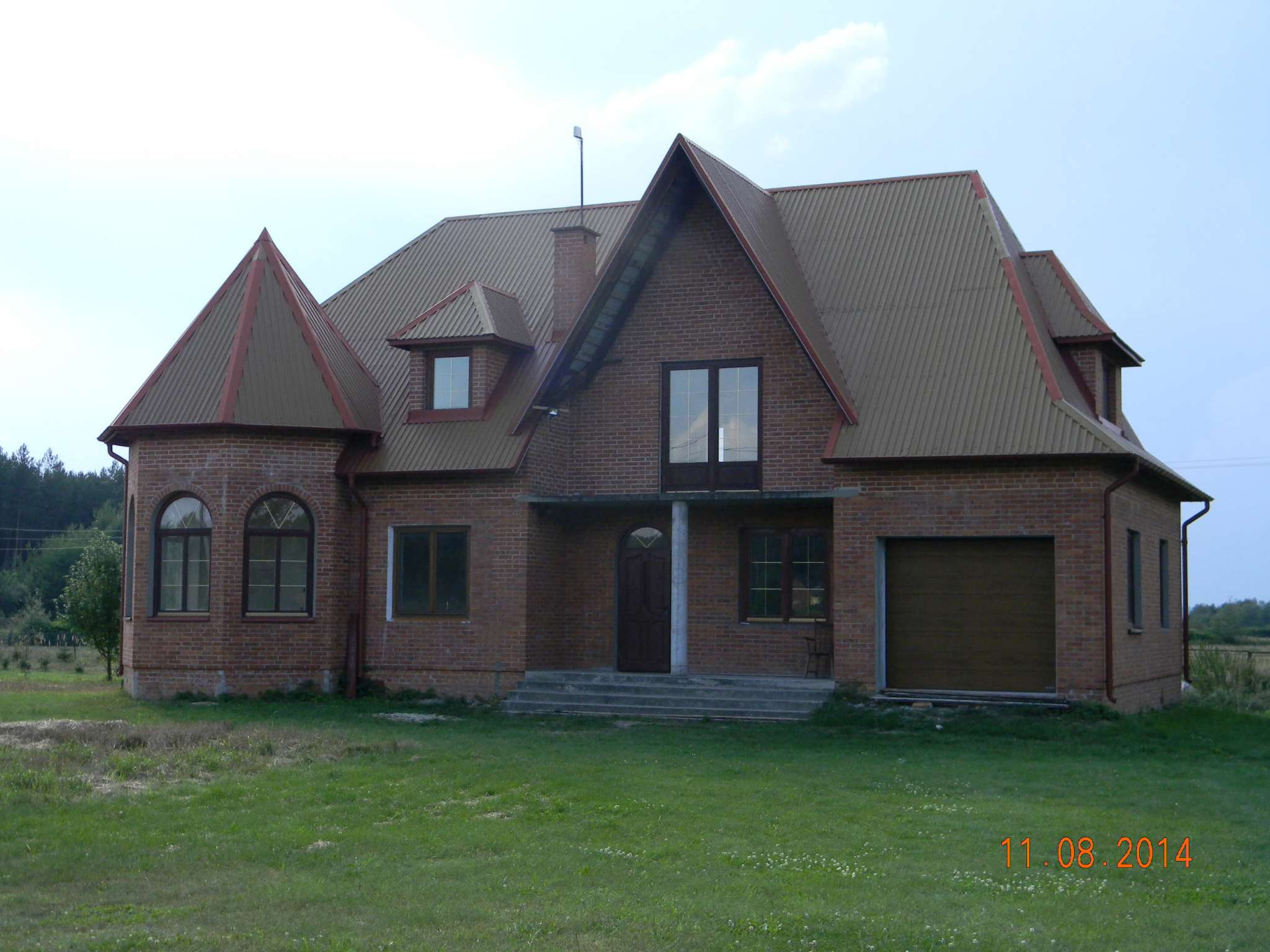 Dwukondygnacyjny dom z czerwonej cegły z brązowym dachem z blachy falistej, widok z przodu na trawnik.