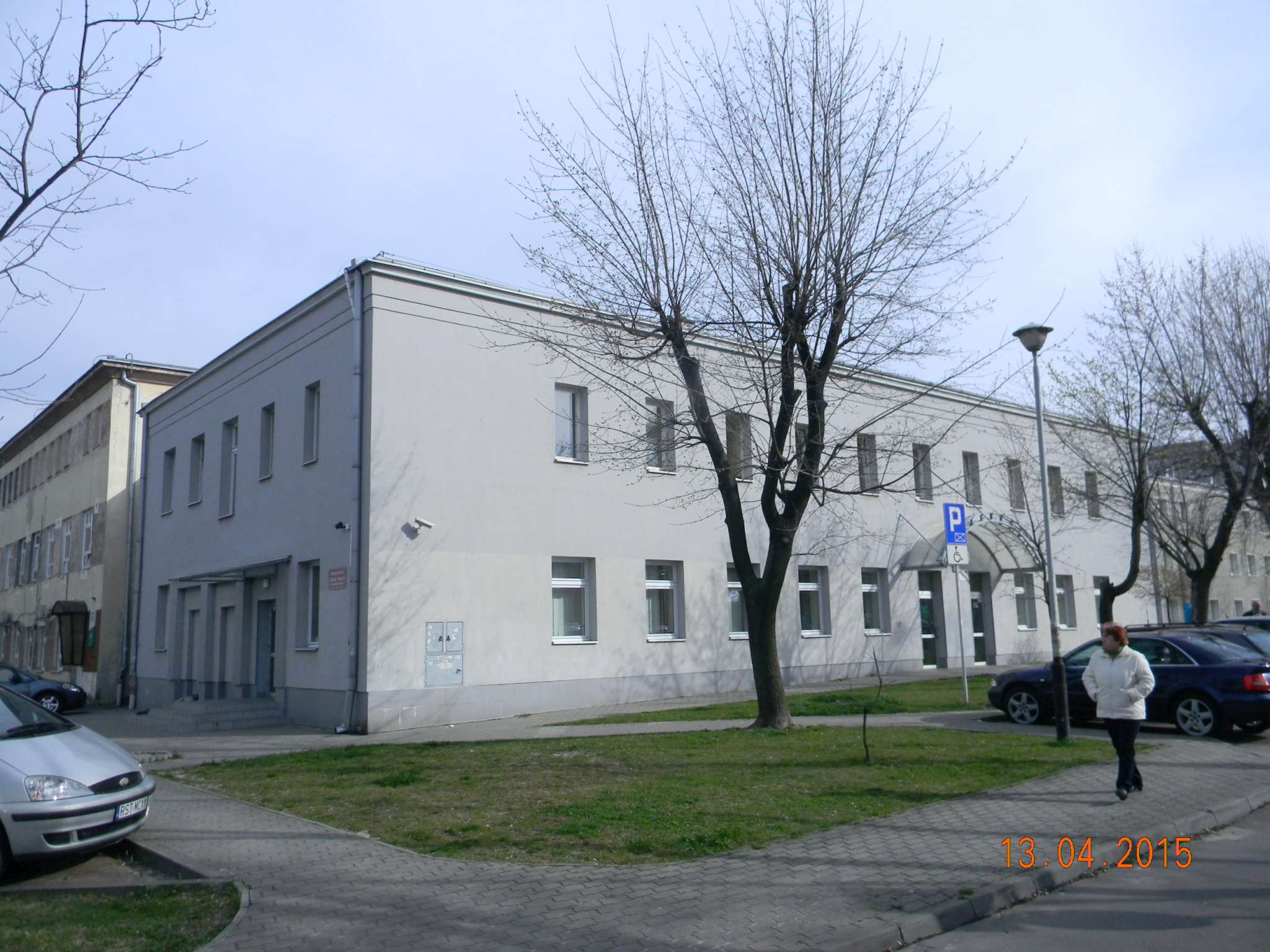 Szary budynek z płaskim dachem i rzędem okien, widoczny znak parkingu dla niepełnosprawnych, drzewa bez liści i chodnik z kostki brukowej.
