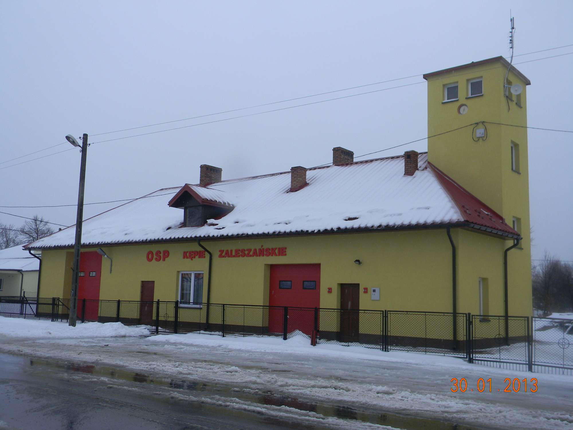 Budynek Ochotniczej Straży Pożarnej w zimowej scenerii, żółta elewacja z czerwonymi bramami garażowymi i dachem pokrytym śniegiem, widoczna wieża obserwacyjna z zegarem.