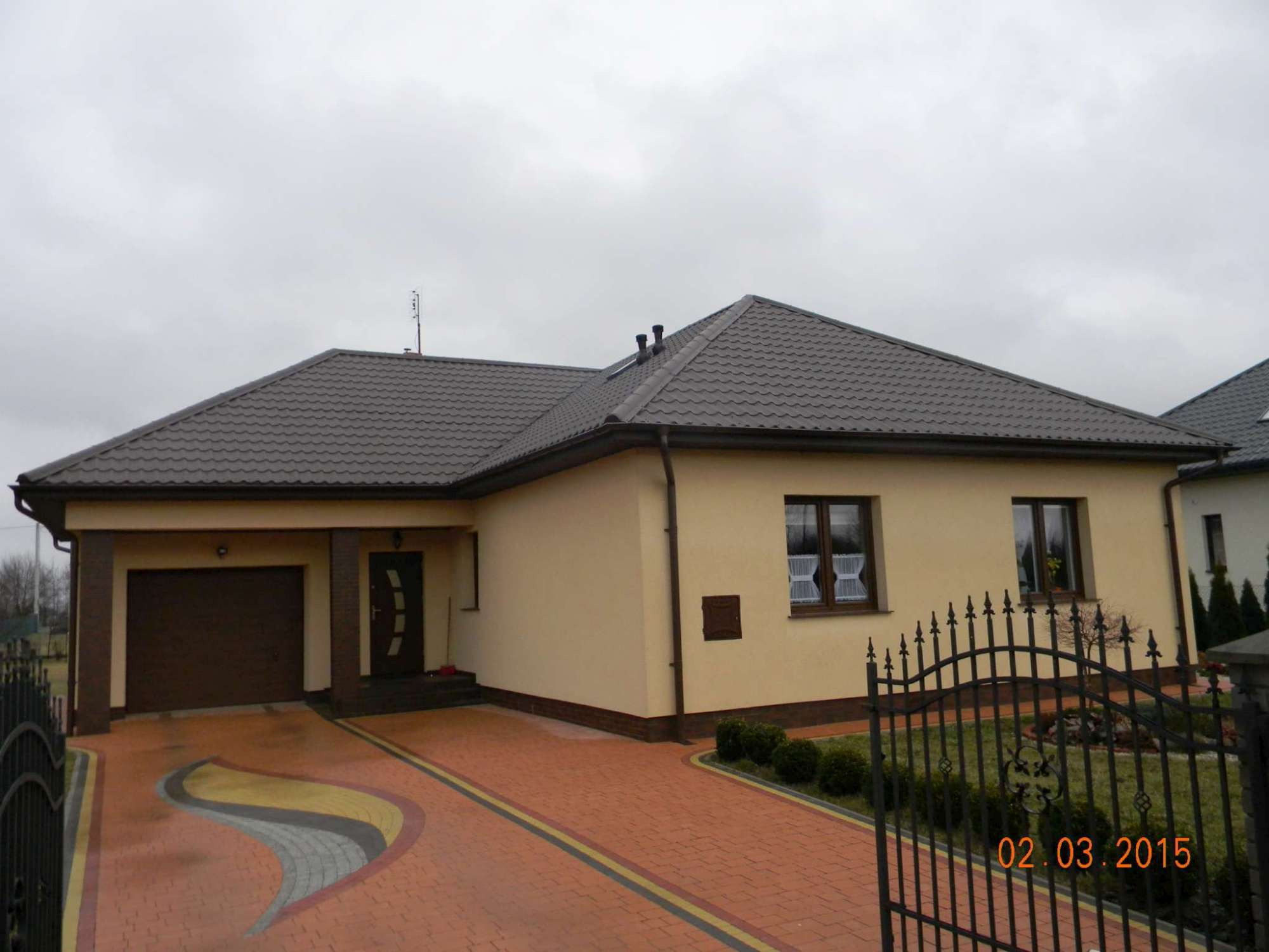 Parterowy dom jednorodzinny z brązowym dachem, beżową elewacją i ozdobnym brukowanym podjazdem, widziany zza metalowego ogrodzenia w pochmurny dzień.