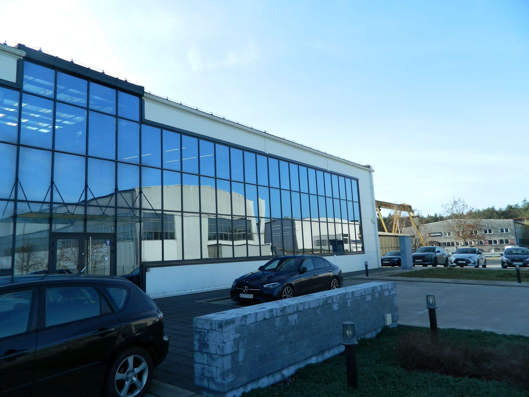 Nowoczesny budynek biurowy z dużą szklaną fasadą odbijającą sąsiedni budynek, zaparkowane samochody na pierwszym planie.