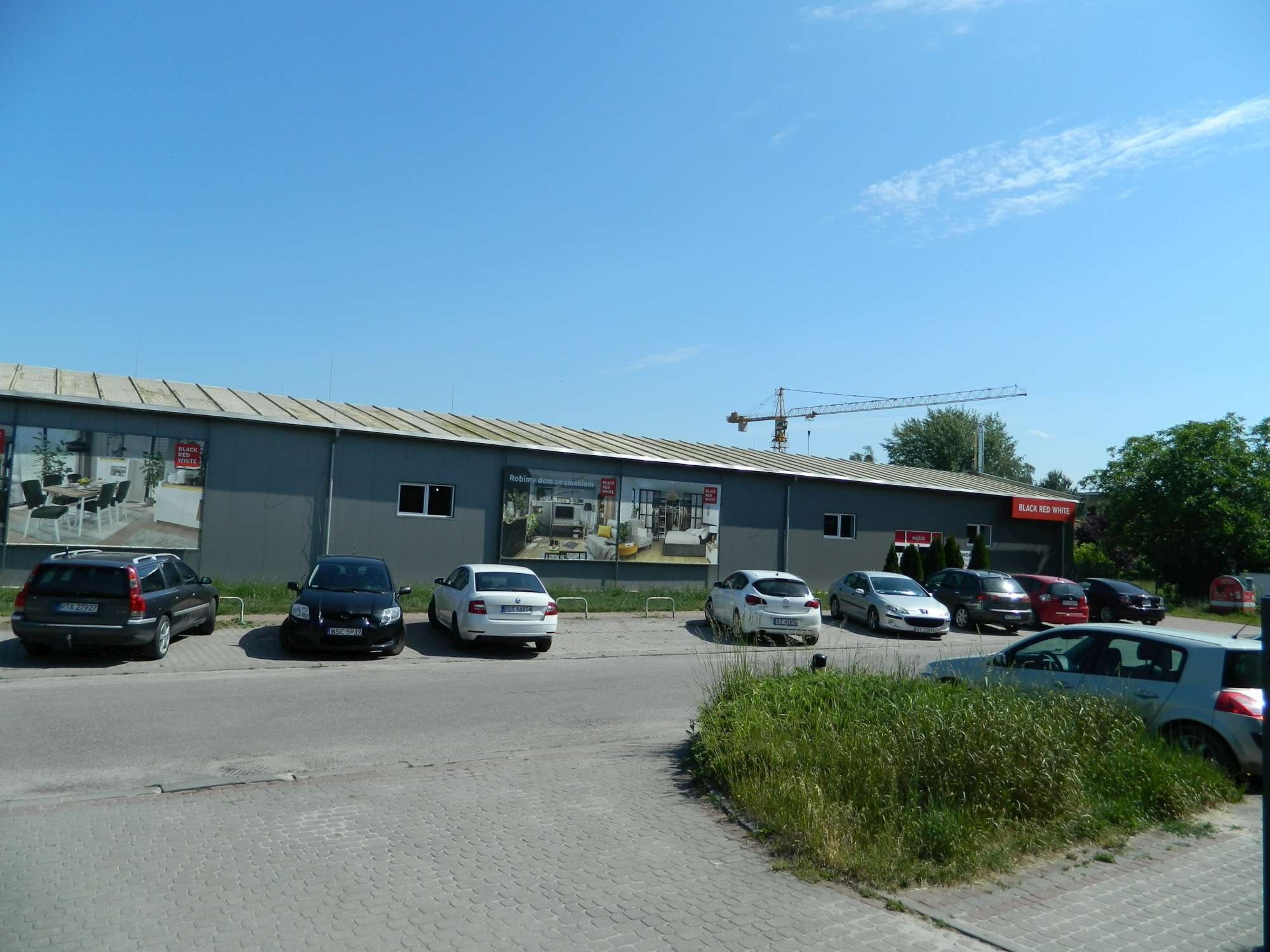 Budynek sklepu meblowego z reklamą zewnętrzną i zaparkowanymi samochodami, na tle błękitnego nieba i żurawia budowlanego.