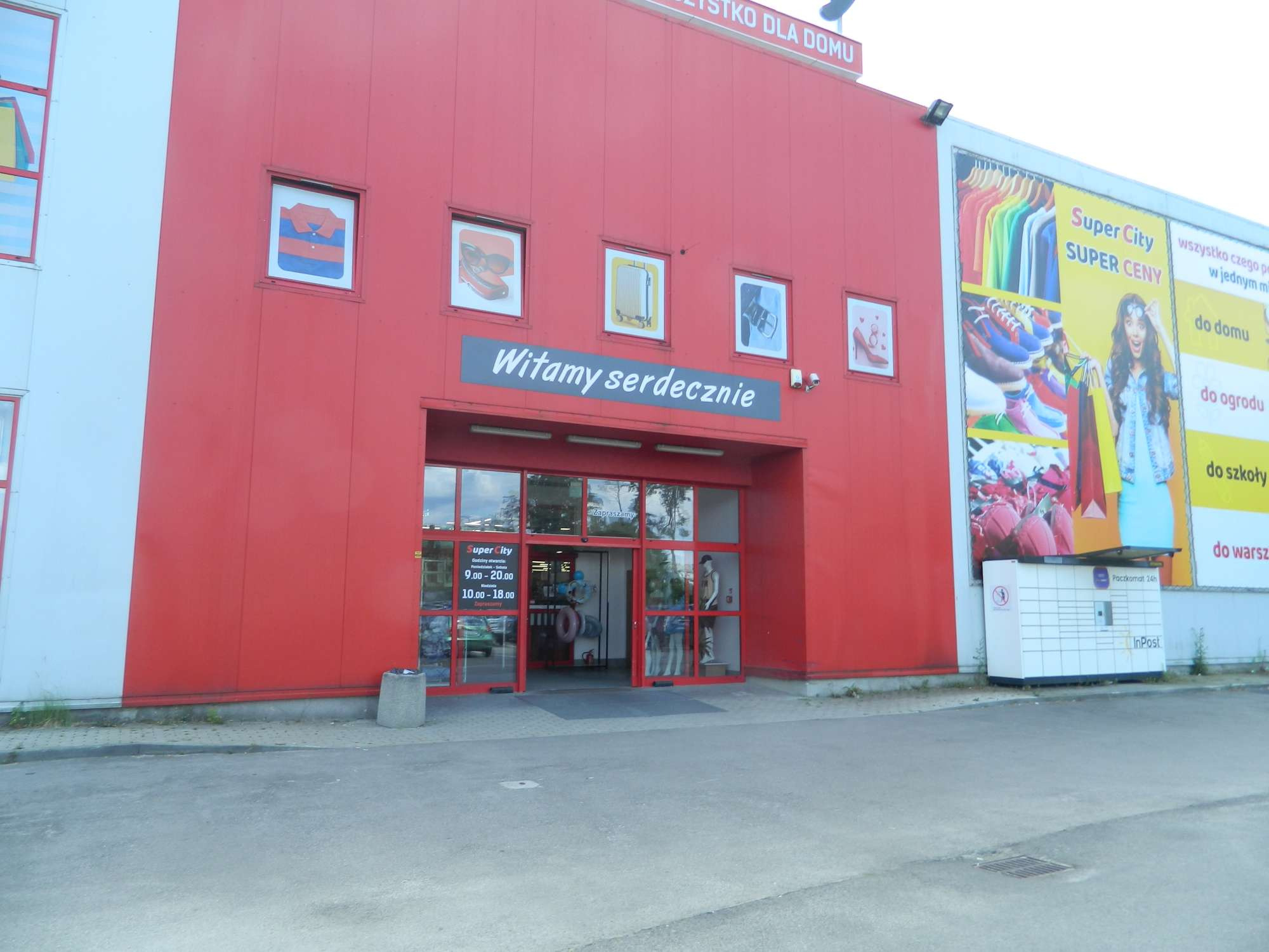 Fasada budynku handlowego Super City z czerwonymi ścianami, oknami z grafikami odzieży i szyldem 'Witamy serdecznie' nad wejściem. Automatyczne drzwi wejściowe i Paczkomat InPost obok.